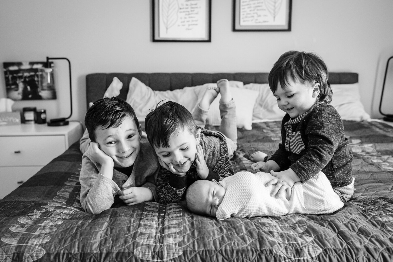 Four young brothers lie on a bed, surrounding their newborn sibling, who is swaddled in a blanket.