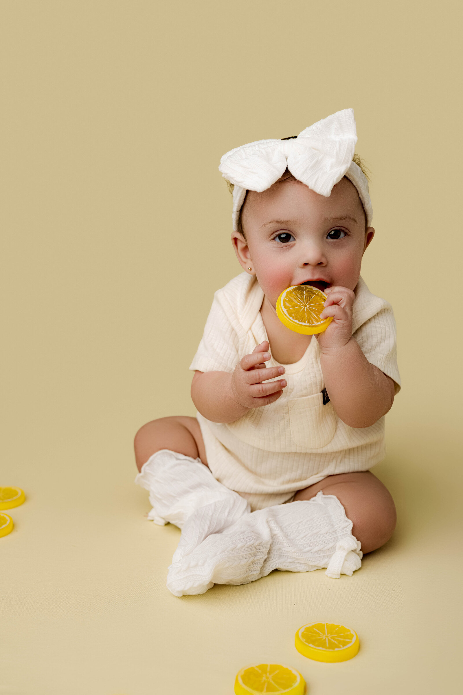 Charlotte baby girl in a white outfit and bow headband, exploring toy lemon slices during her milestone photography session.