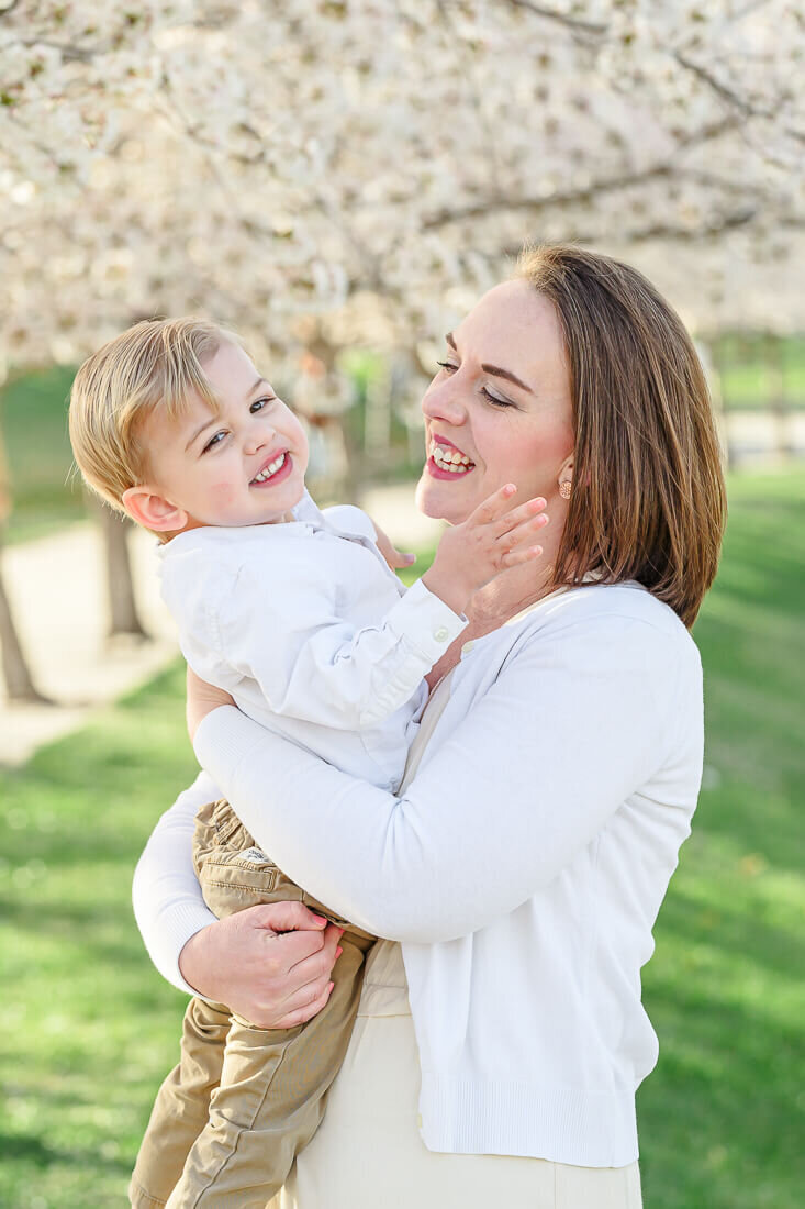 A mother holds her toddler son and smiles at him while standing in front of blossoming cherry trees on the grounds of the Utah State Capitol. Captured by Salt Lake photographer Melissa Woodruff Photography.