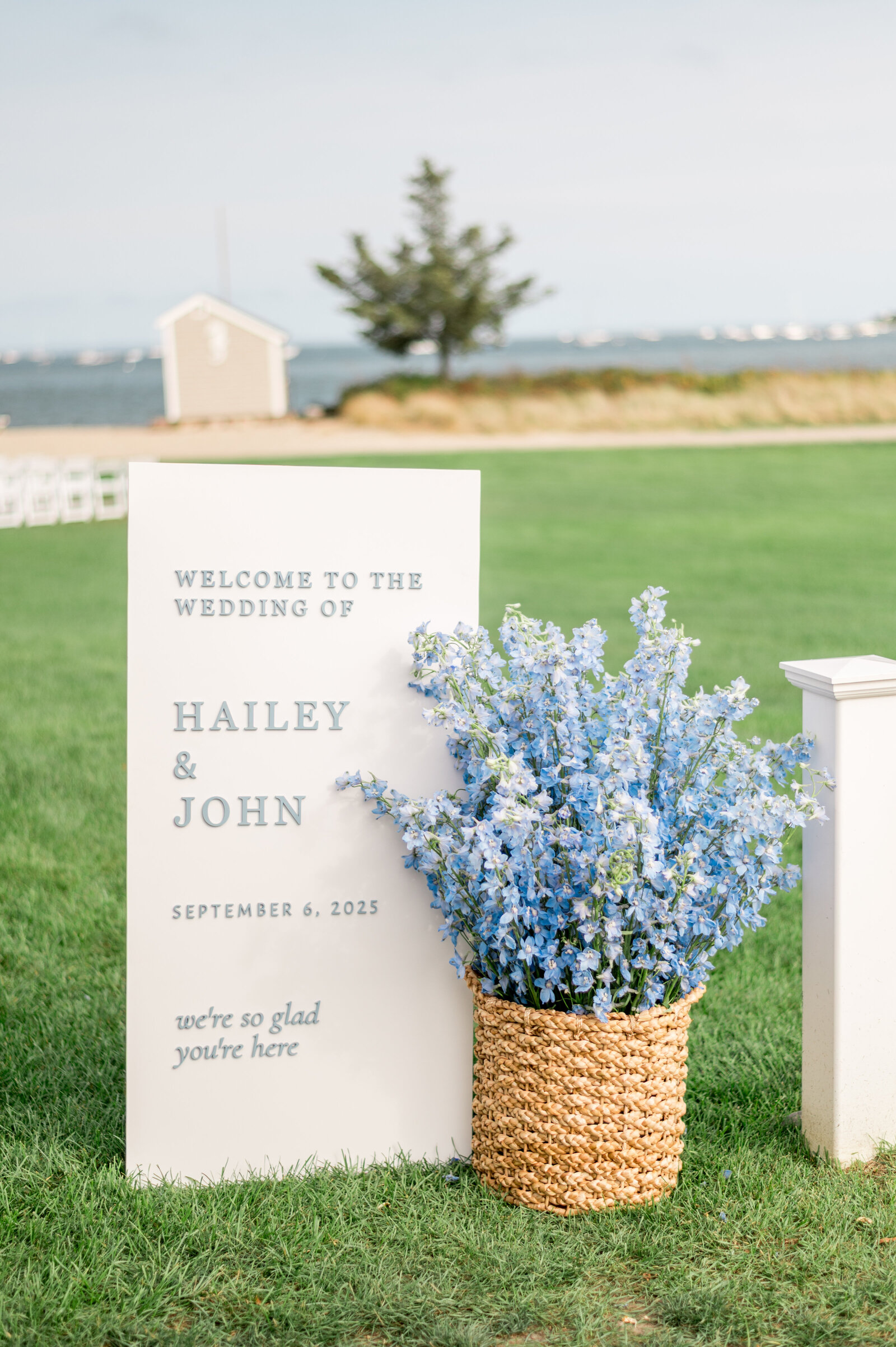 Elegant white wedding welcome sign with blue floral arrangement at coastal New England venue — detail photography by Sarah Surette Photography.