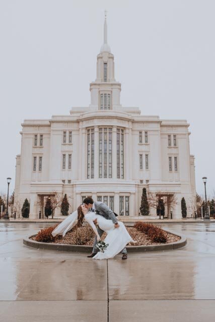 groom dipping his bride and kissing her in front of an LDS temple on a rainy day