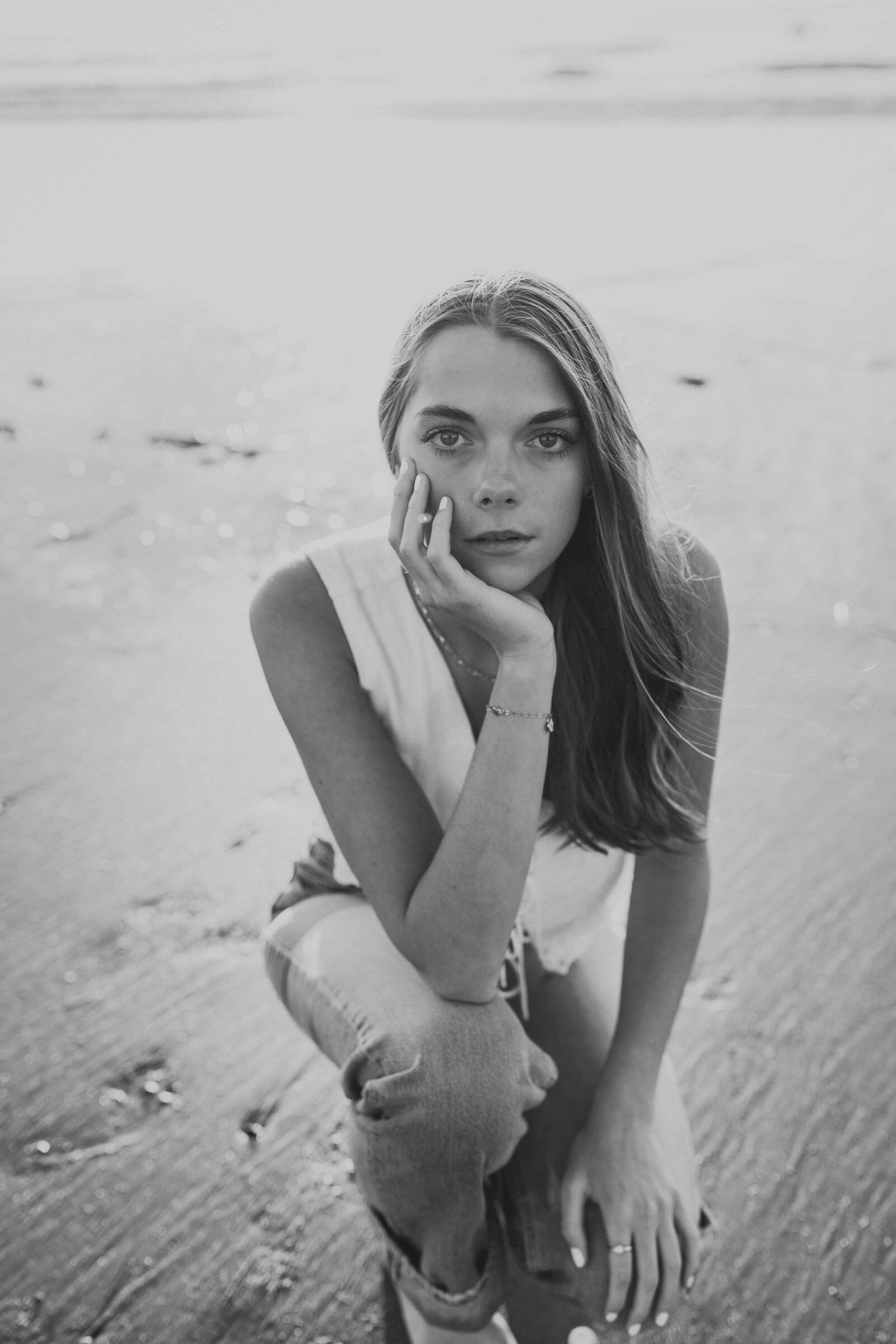 black and white photo of brunette high schooler posing on a beach wearing a white shirt