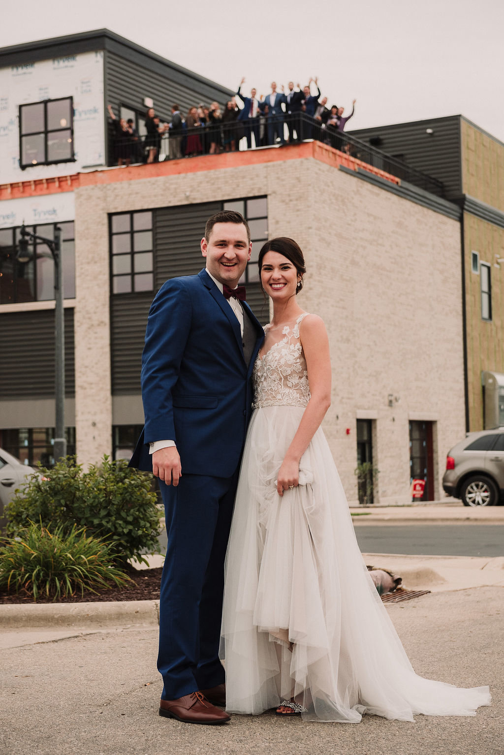 Bride and groom posing on the ground with party on the rooftop