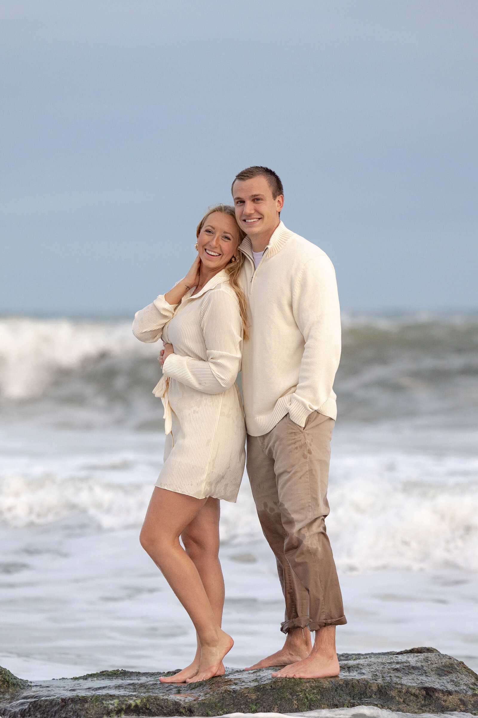 A couple standing confidently on coquina rocks by the ocean in Fort Fisher, NC, with waves crashing behind them. This elegant wedding photography highlights their connection, appealing to couples looking for professional wedding photography packages in NC.