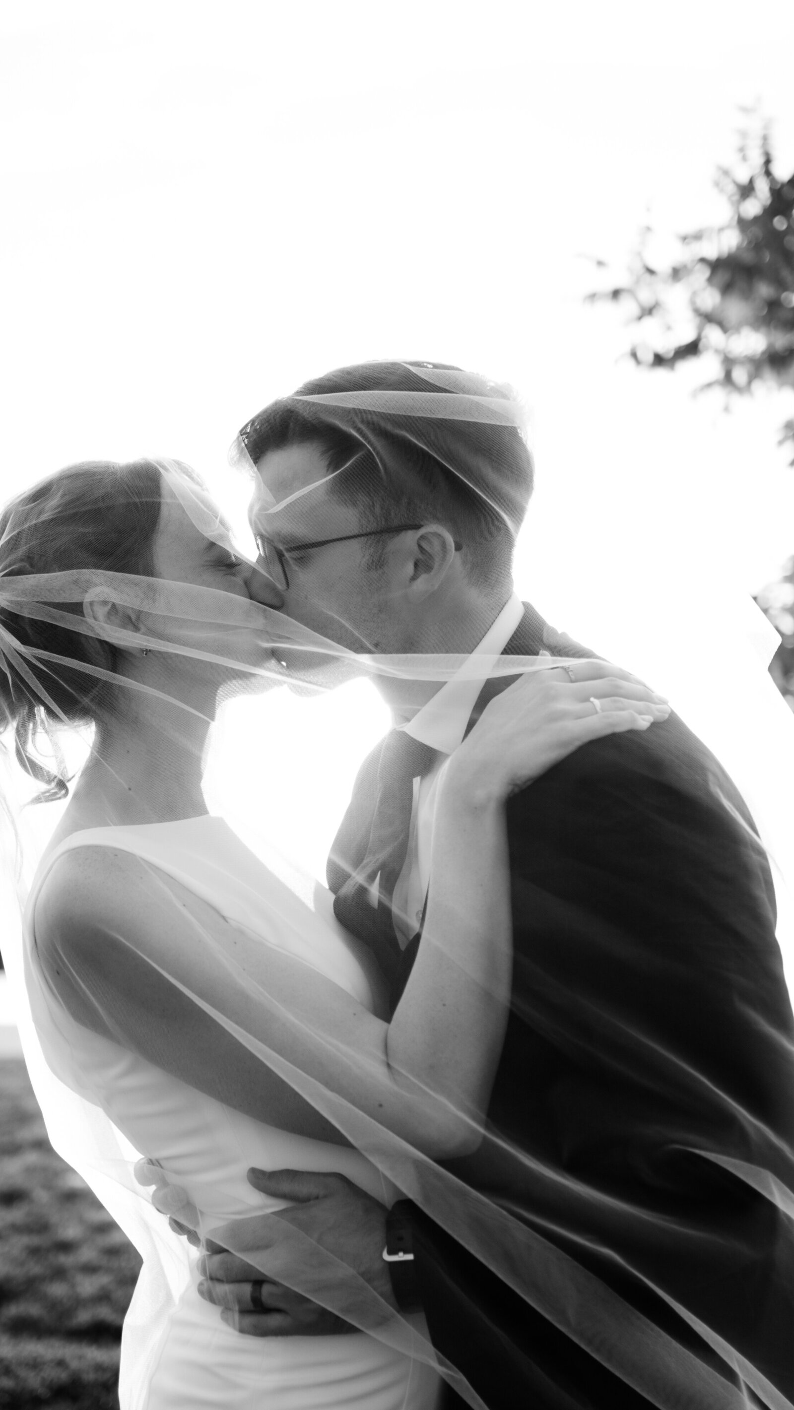 Couple under veil kissing. Her hands are on his shoulders. 