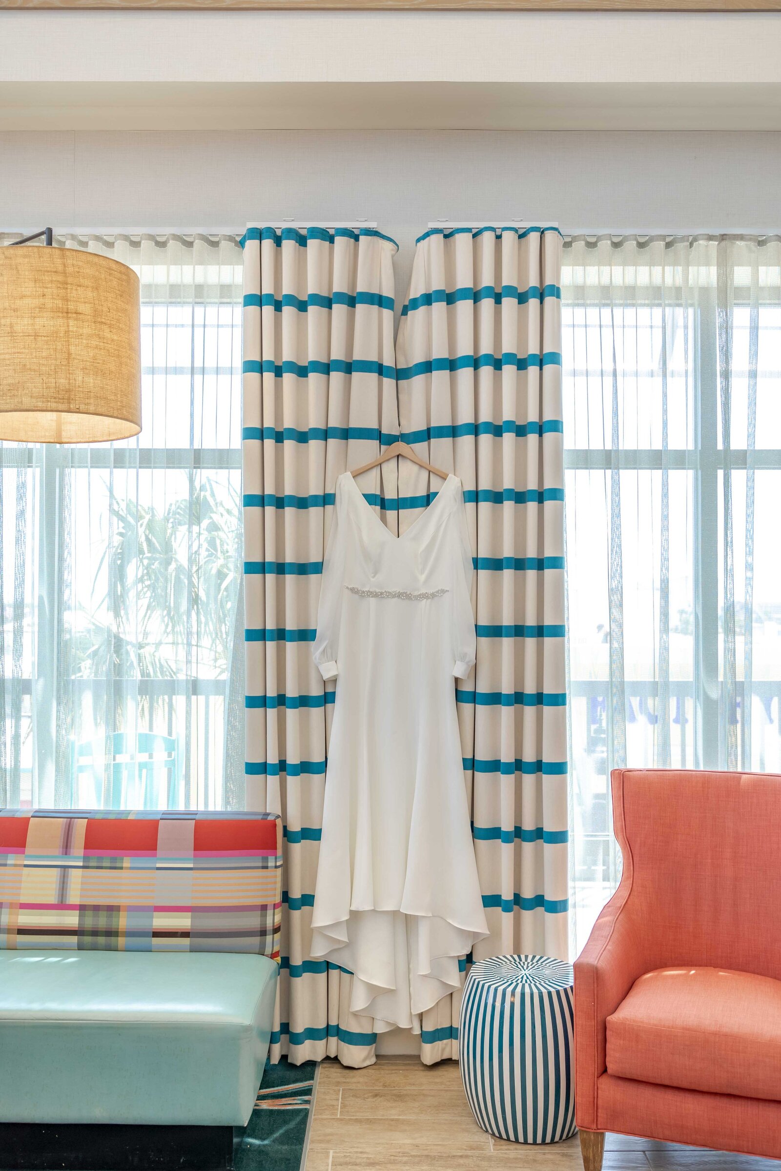 A simple yet elegant wedding dress hanging against striped curtains in a colorful and modern bridal suite, captured during a Wrightsville Beach, NC wedding, blending contemporary style with coastal charm.