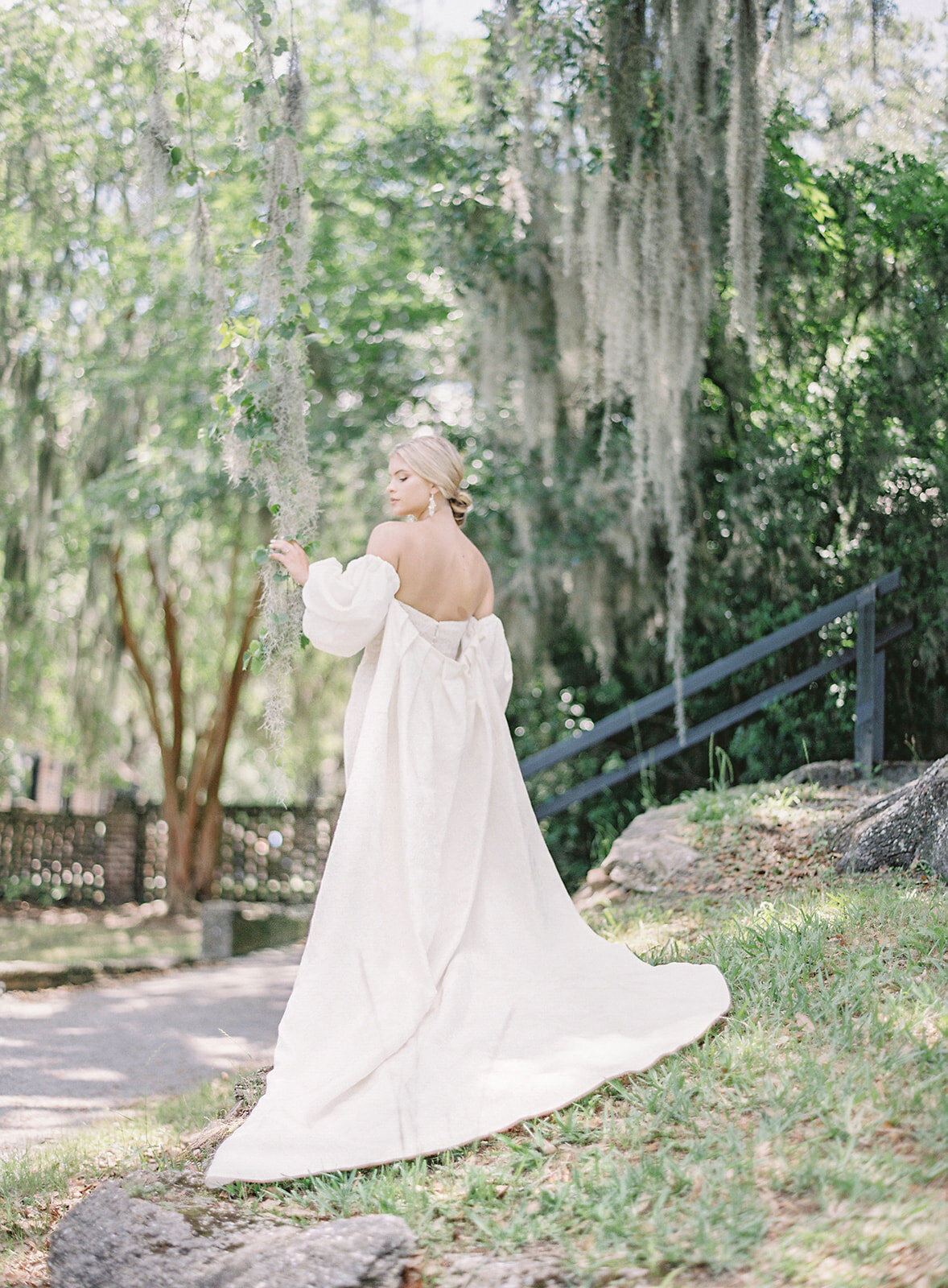 Bride standing on small hill holding on to spanish moss hanging from an oak tree. She is facing away from the camera and looking to her left so that her profile is shown. She has a low chignon bun and is wearing an ivory strapless wedding gown and an ivory cape with train that shows her shoulders and back with puffed sleeves designed by Anne Barge. Photographed by wedding photographers in Charleston Amy Mulder Photography.