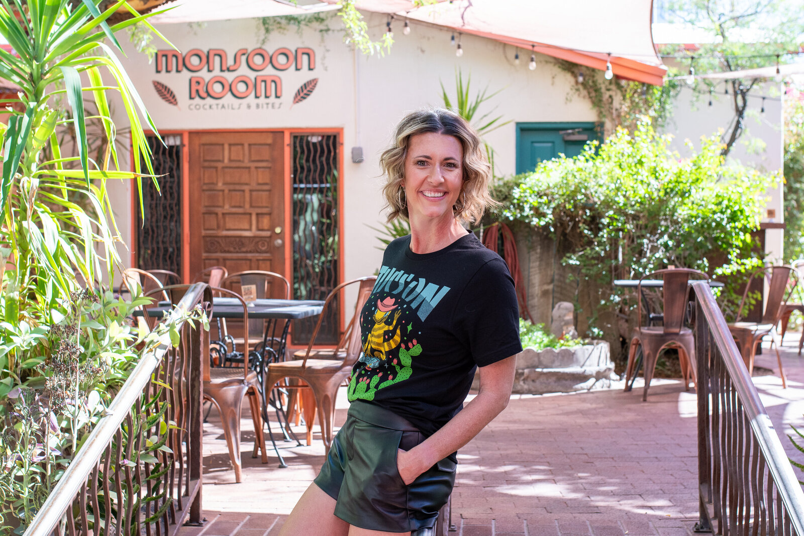 Smiling woman standing outdoors near a café entrance with plants and patio seating, photographed by Vyrl Photo for Tucson brand photography.
