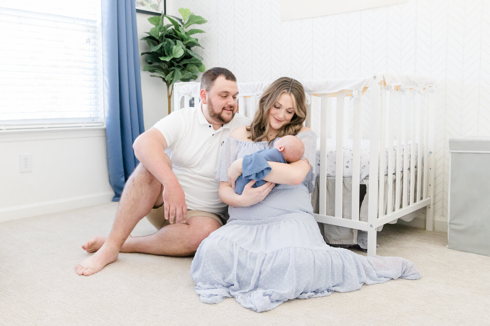 Mom and dad sit on floor in front of crib while holding baby and smile down at him during newborn session