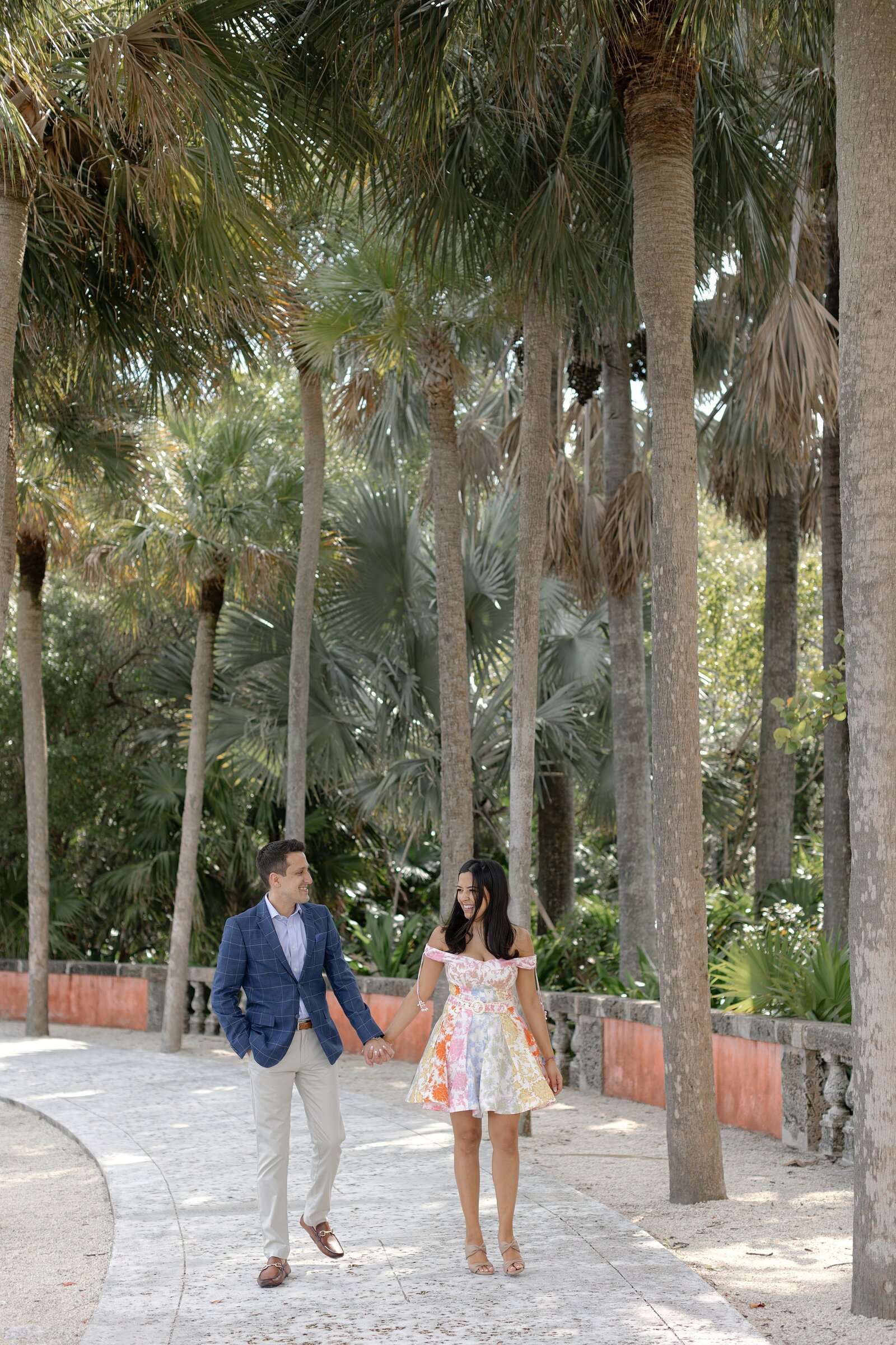 Couple walk hand in hand towards camera on a palm tree lined walkway at Vizcaya Miami.