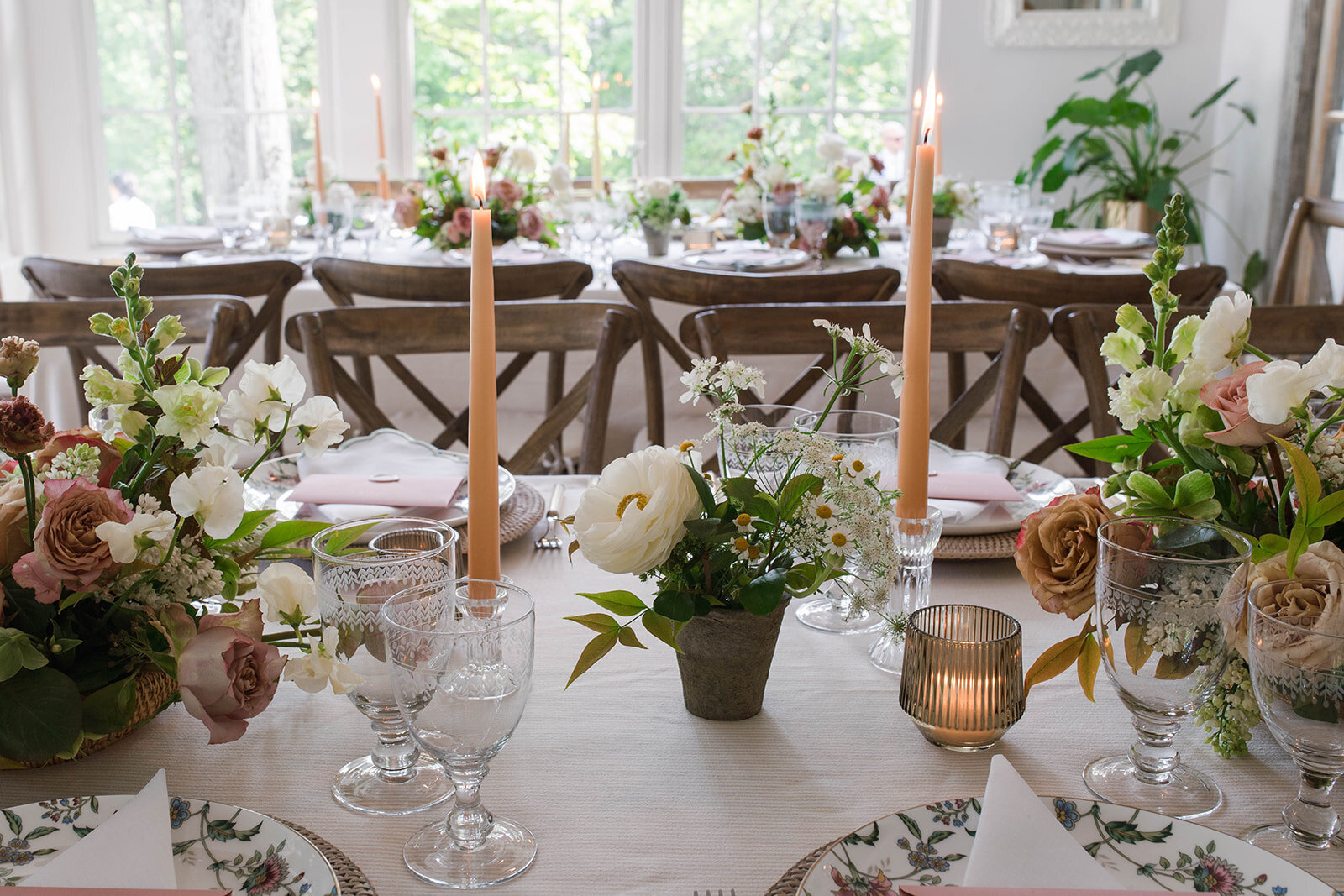 A long table with floral arrangements including white ranunculus, white snapdragons, brown garden roses, white sweet pea, daisies, mauve garden roses, and greenery with sand-stone colored taper candles, amber ribbed votives and vintage glass-ware.
