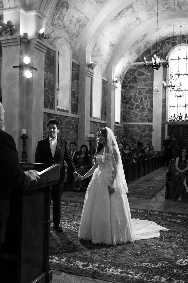 A bride and groom stand holding hands before a minister in an ornate church, with stone walls and a high, decorated ceiling. The scene is black and white, and guests are seated in pews, observing the ceremony.