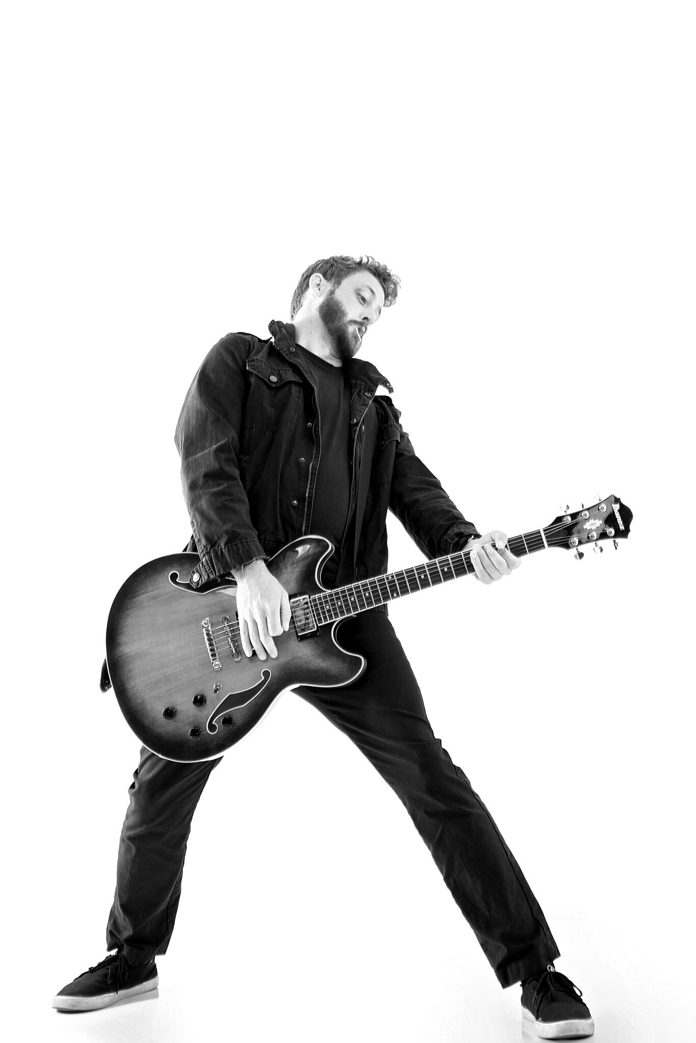 A man in dark clothing stands with legs apart, playing an electric guitar. The white background creates a high-contrast, dramatic effect—ideal for band photography Jacksonville professionals seek.