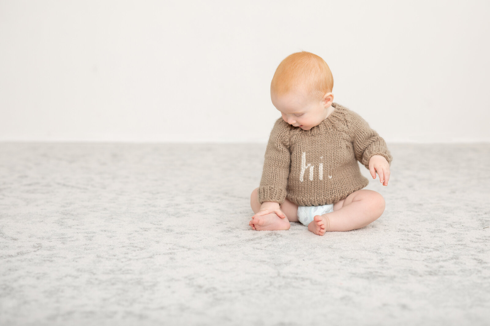 Toddler wearing a brown sweater with the word 'hi' sits on the floor, smiling softly. Captured by Bucks County Photographer Ivory Tree Portraits