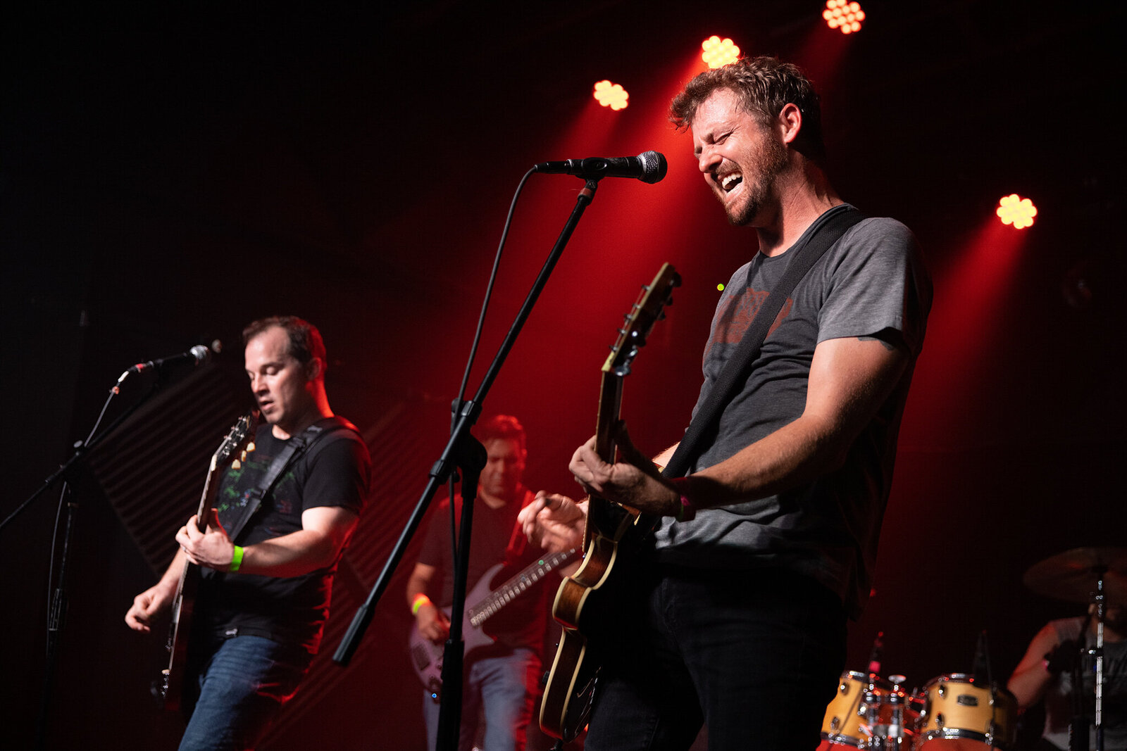 A rock band performs on stage under red lights; two guitarists are at the front, one singing passionately into a microphone, while a bassist and drummer play in the background—perfect for Concert photography Jacksonville.