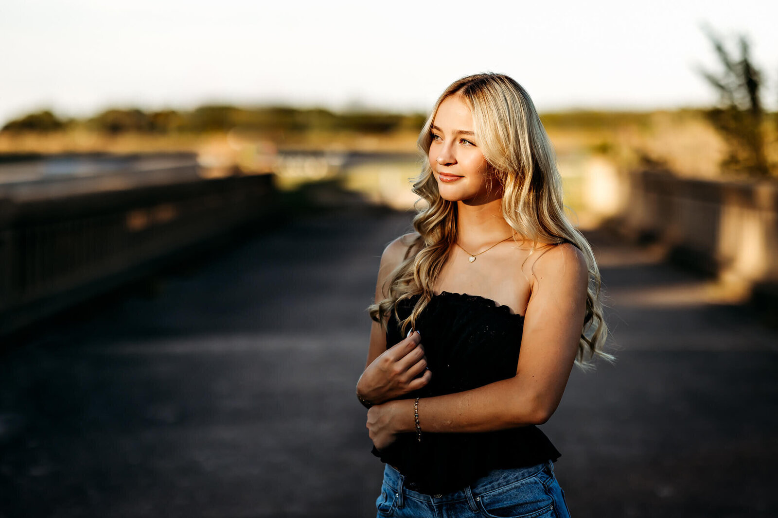 A young woman with long blonde hair stands outdoors, wearing a black top and blue jeans. She is looking to the side with a slight smile. The background is blurred, with a bridge and greenery visible in the distance. The lighting is warm and golden.