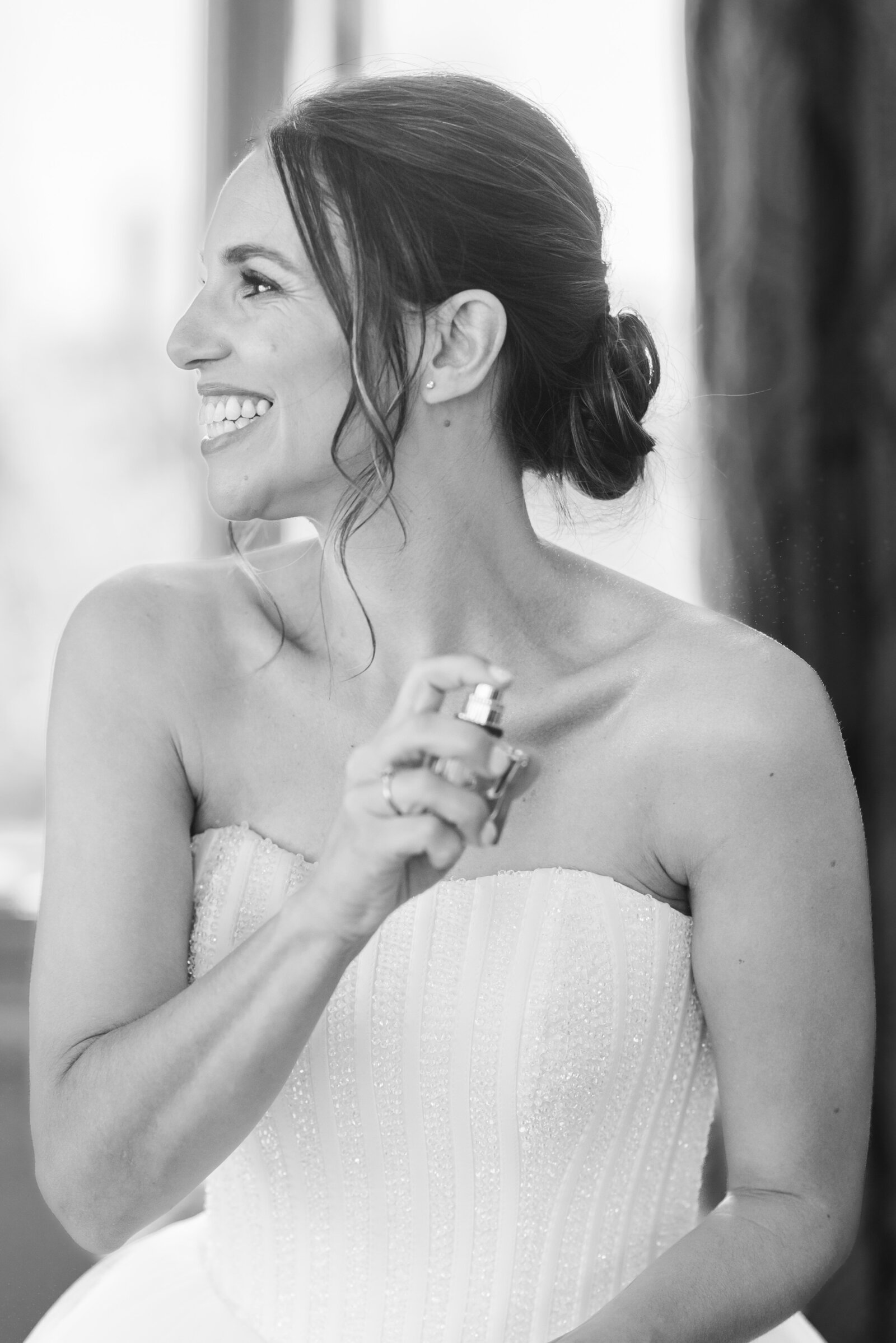 Black and White image of a bride spraying on her perfume, she is looking to the left and smiling. Her dress is strapless and ballgown in style.