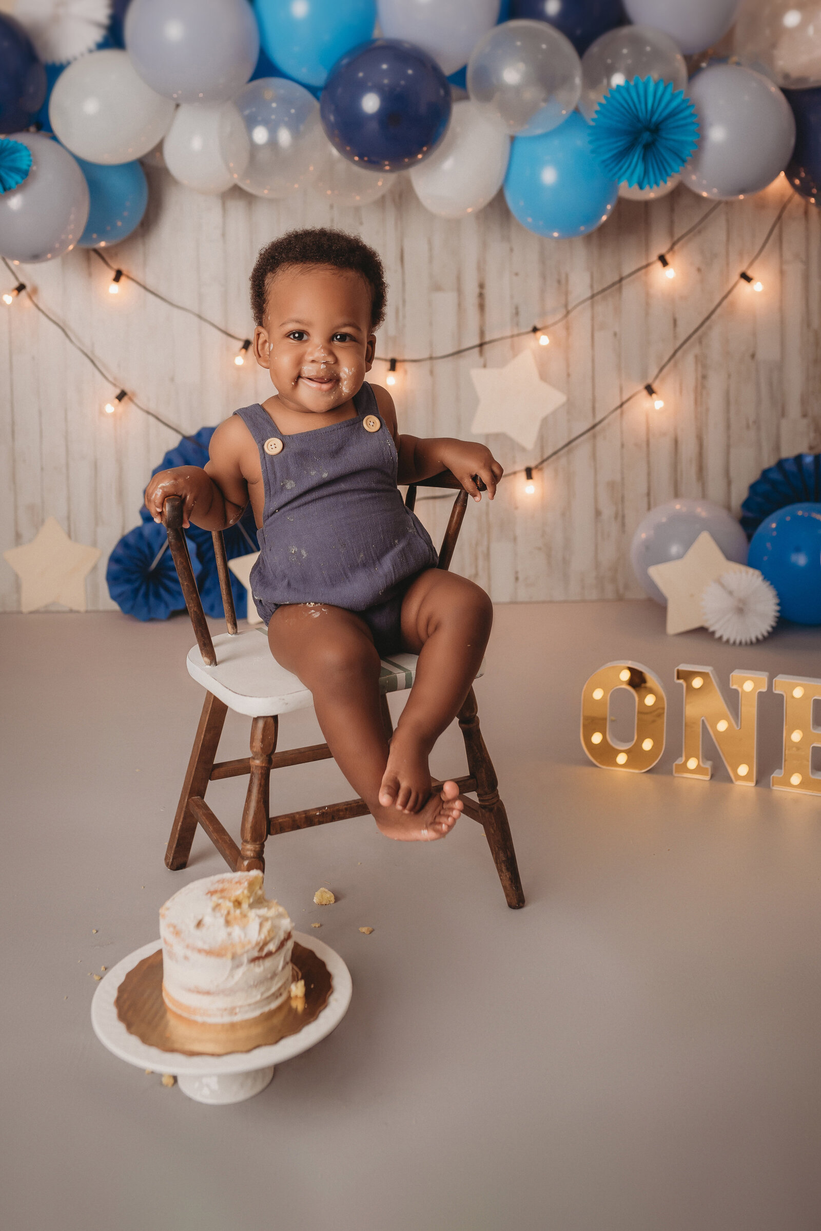 one yar old baby boy at Marietta GA cake smash session sitting in chair wearing blue romper with smash cake beside him and blue balloon garland with string lights in background