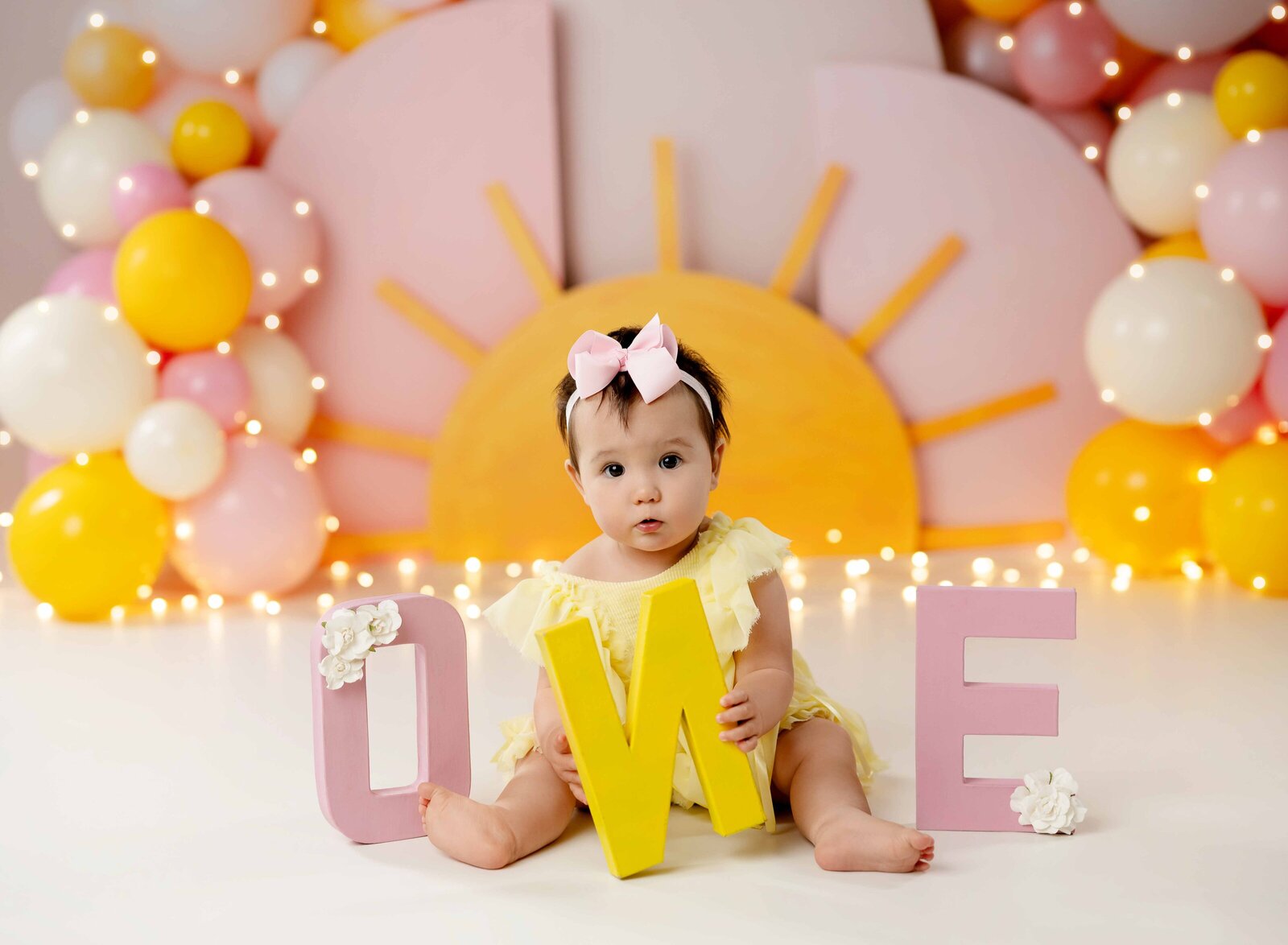 Baby on shine backdrop with pink and yellow balloons, holding ONE letters and a cute look on her face.