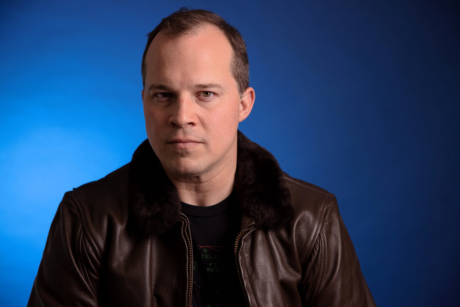 A man with short brown hair wearing a dark brown leather jacket over a black shirt poses against a blue background, looking directly at the camera with a neutral expression—a great example of professional band photos Jacksonville style.