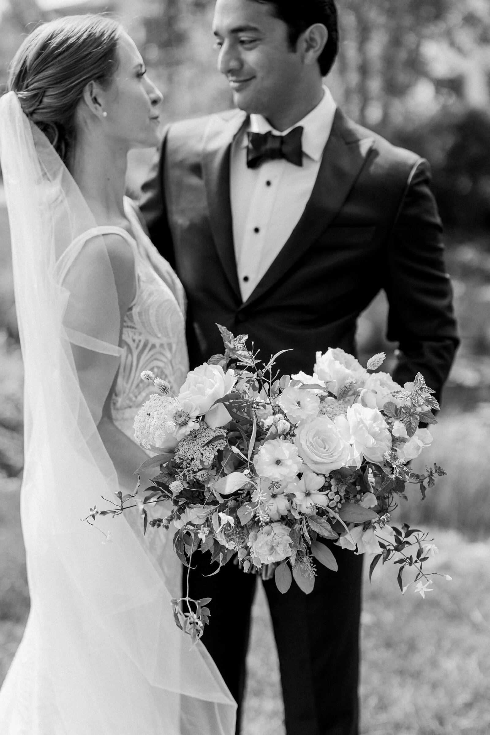 Black and white photo of a wedding couple smiling at each other while one holds a bouquet of flowers.