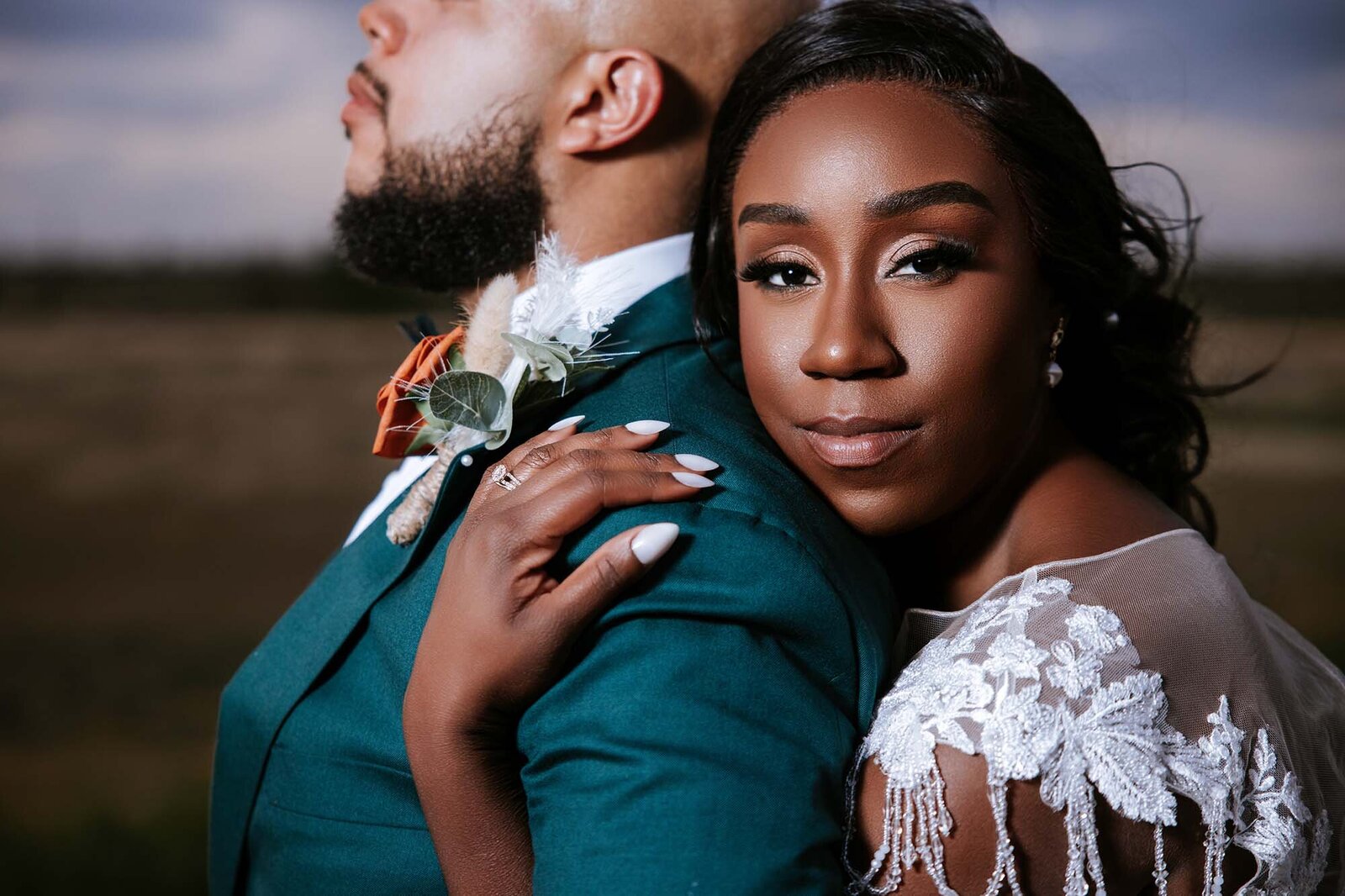 Elegant bridal portrait captured by Houston wedding photographer Logrono & Co., featuring an affectionate embrace between an African-American bride in a lace gown and her groom in a green suit with a rustic boutonnière.