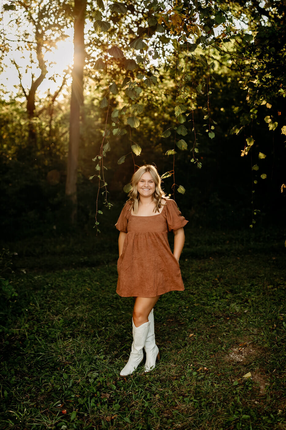 A woman stands on a grassy area in a forest. She's wearing a brown dress and white boots. The sun shines through the trees in the background, creating a natural, serene atmosphere.