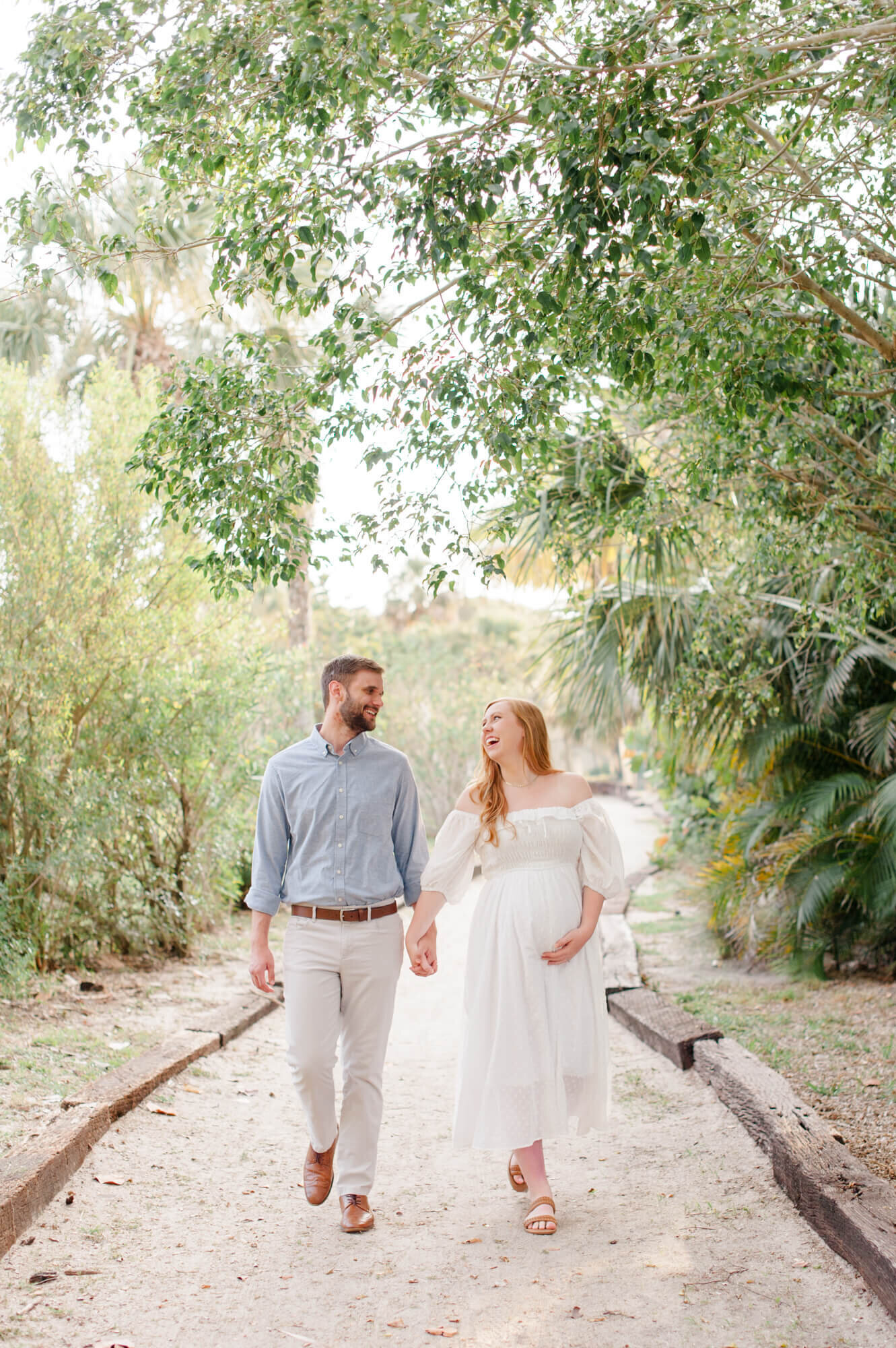 Pregnant mom holds her husbands hand and walks on a beautiful greenery beach path during their maternity photoshoot