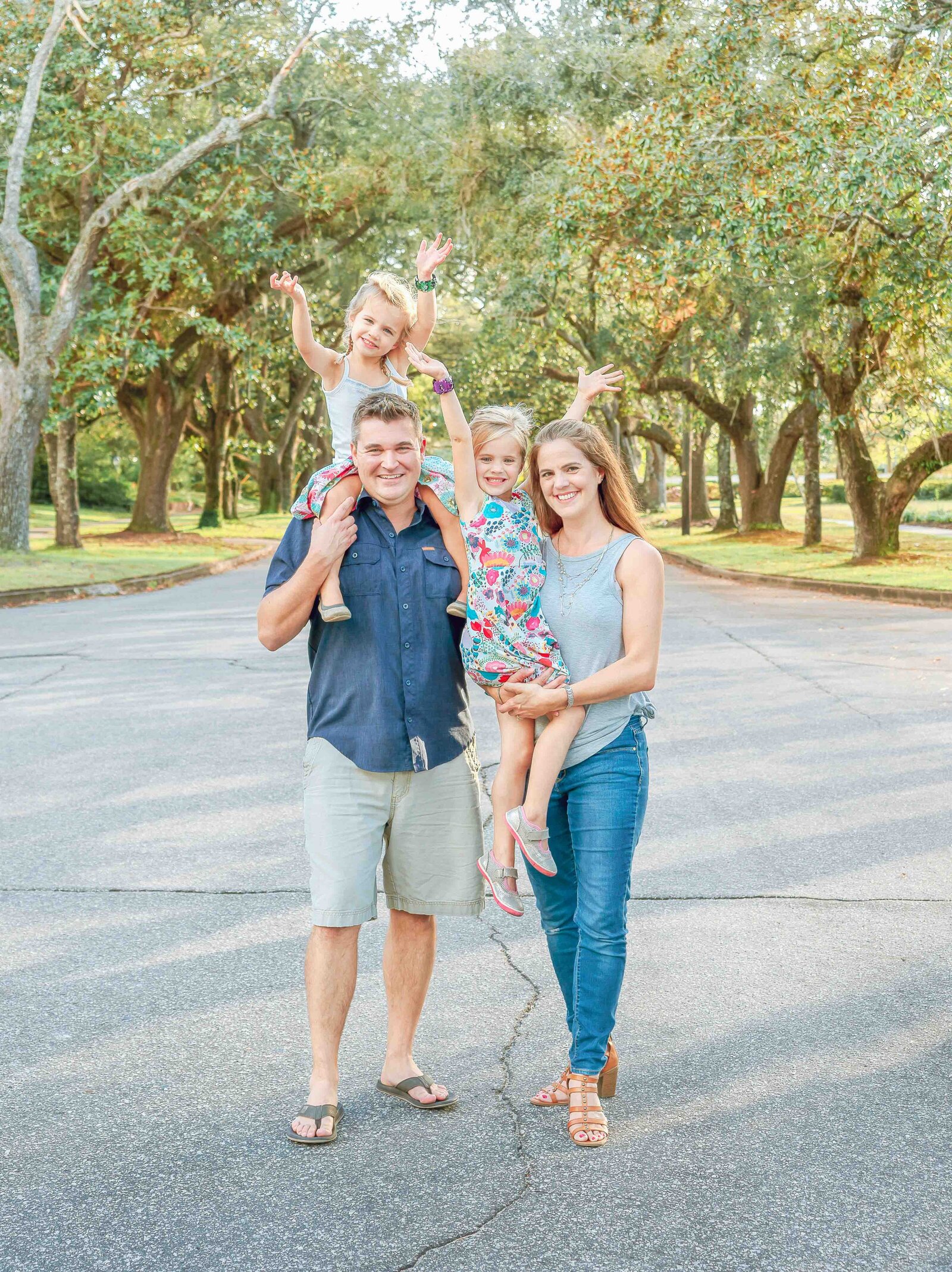 A family stands in a wooded park, with two young children playfully perched on their parents' shoulders. Dressed in casual summer outfits, they share joyful smiles. This image highlights artistic family photography in Wilmington, NC, capturing lively and candid moments.