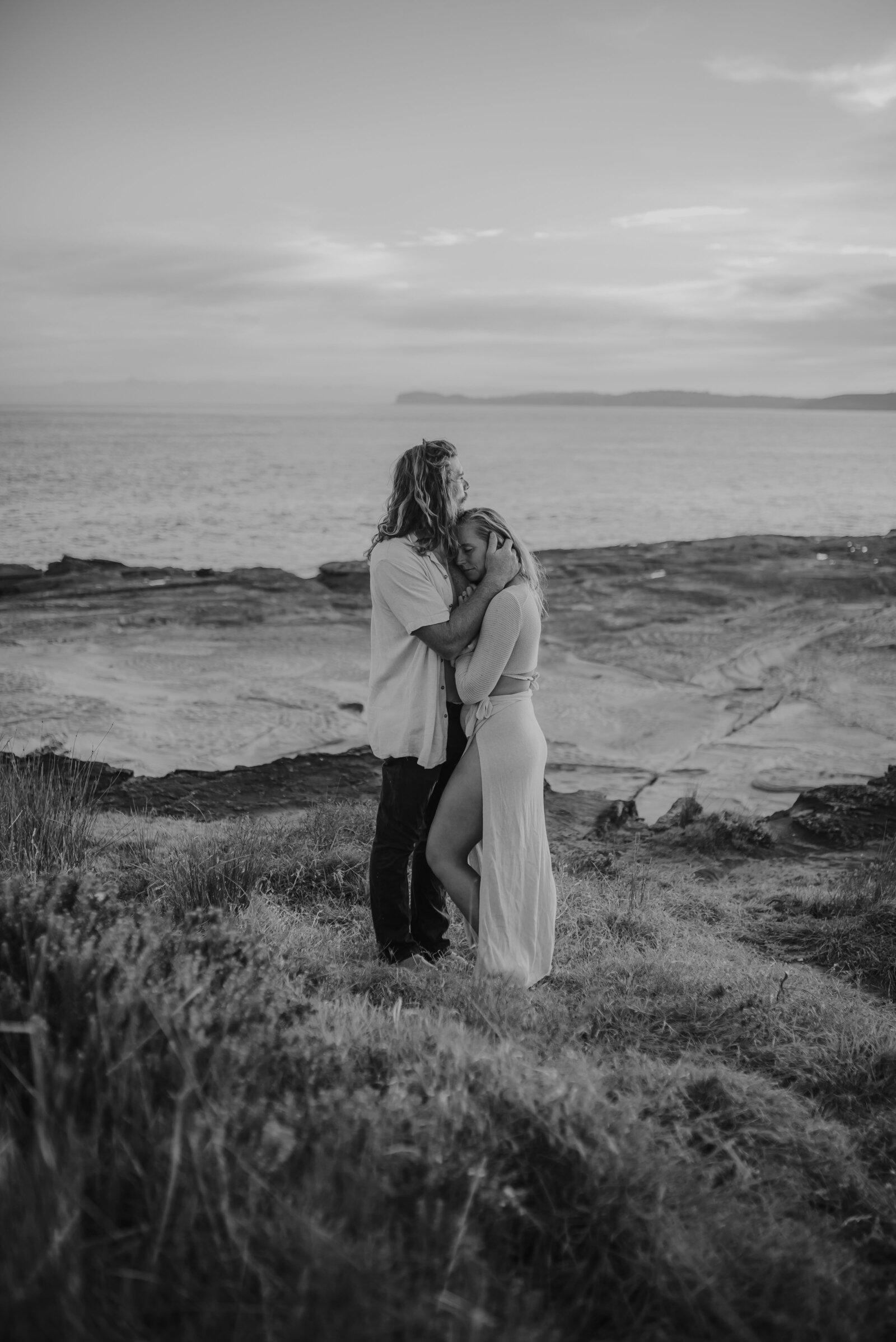 A couple stands in an embrace on a grassy cliff overlooking the ocean, with the man kissing the woman's forehead. The monochrome image captures a serene moment at dusk.