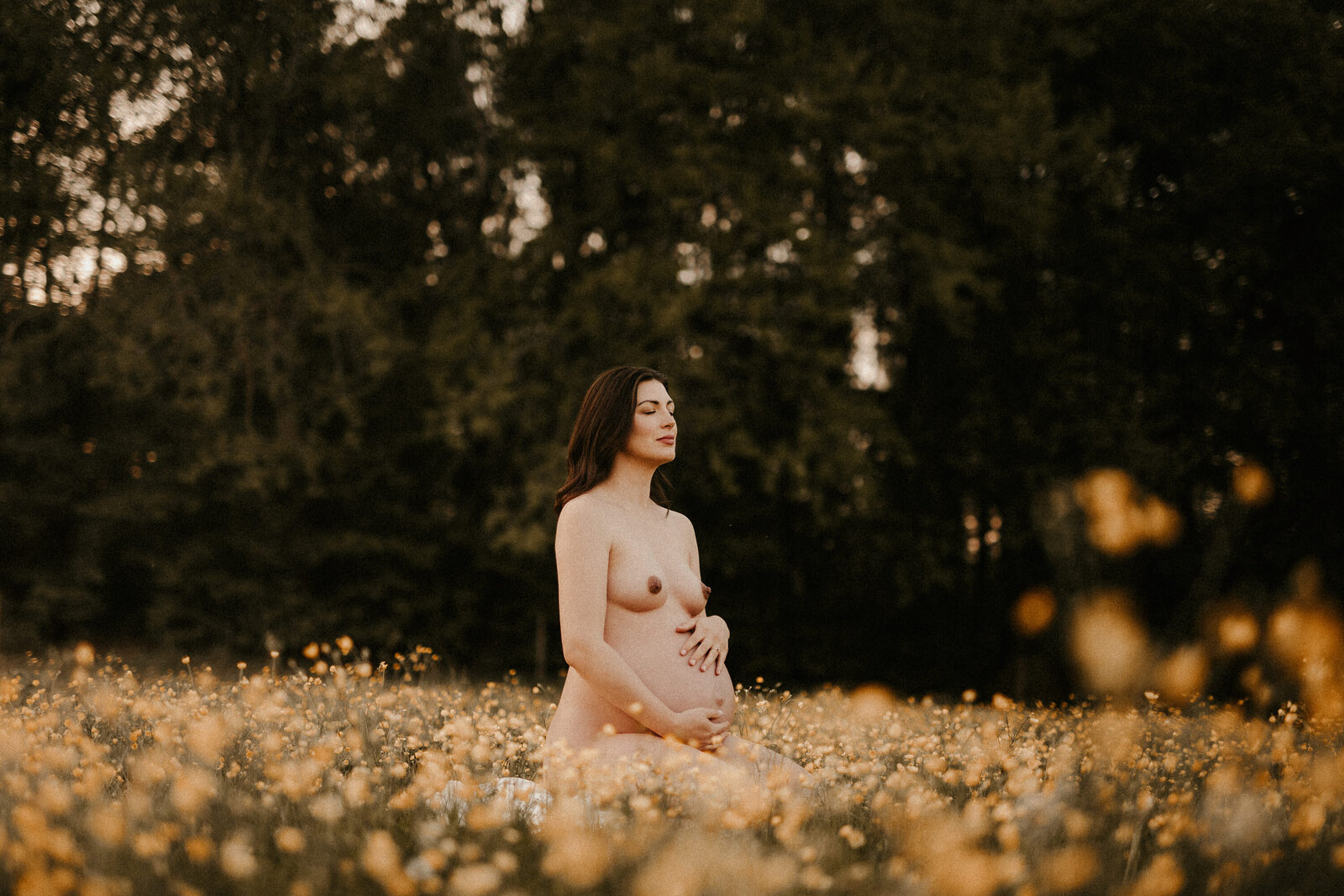 en naken gravid kvinna sitter på ett blomfält och håller om sin mage med en stolt blick