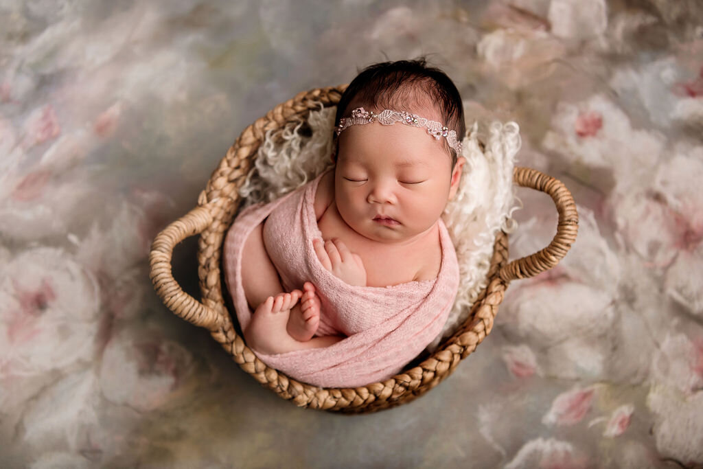 Professional Connecticut newborn photographer showcases baby in blush pink wrap and delicate headband in wicker basket