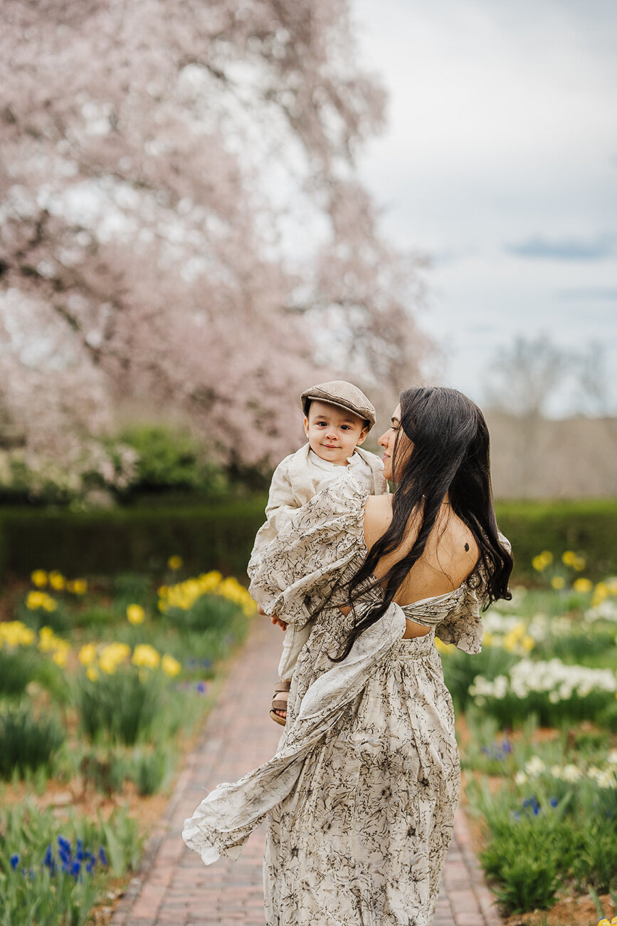 mother with long dark hair holds baby son while dress blows in spring garden
