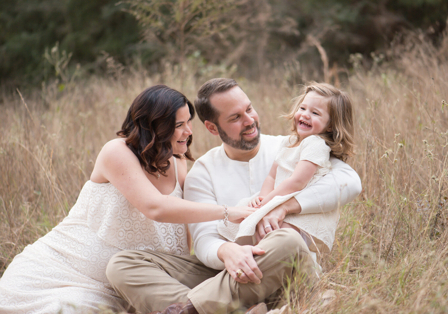 Mom, dad, and daughter share a joyful moment in a tall grassy field. A golden-hour family session by Moments in Light Photography.