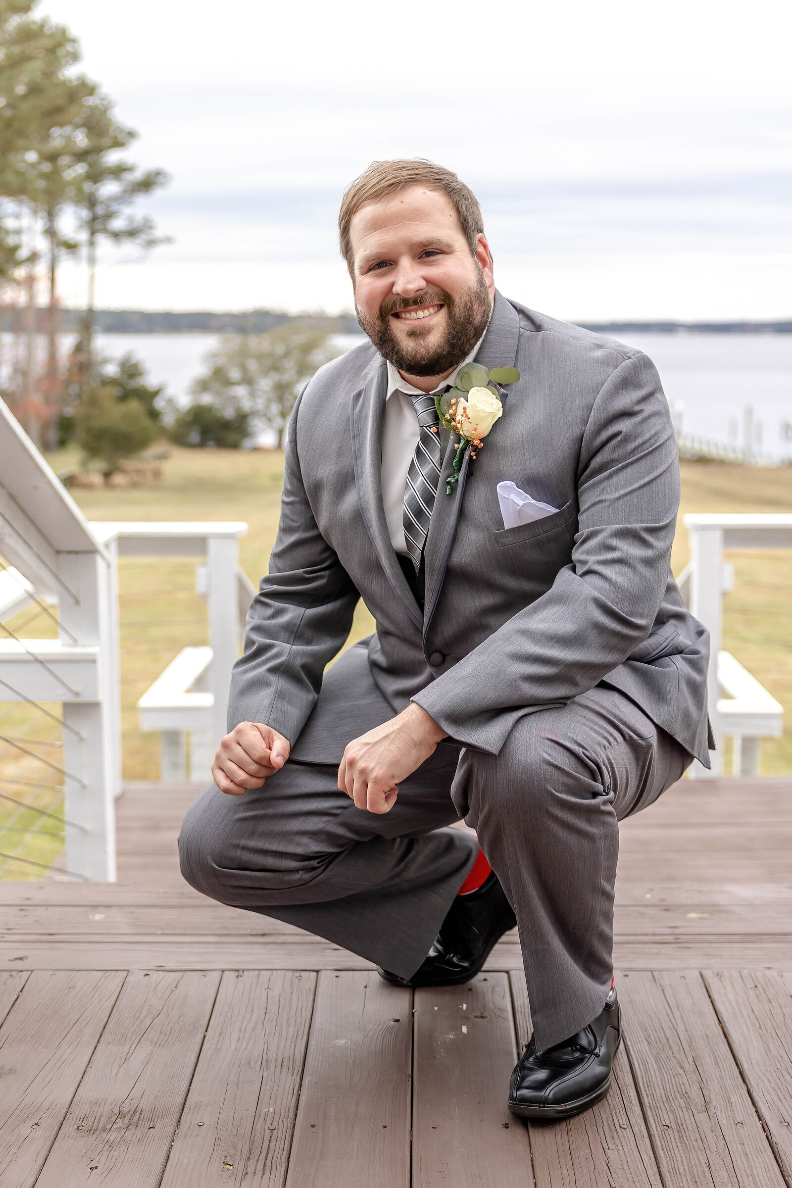A groom poses on a wooden deck with a scenic water view in the background, wearing a gray suit and boutonniere, captured at a Pine Knoll Shores, NC wedding, showcasing classic elegance and waterfront charm.