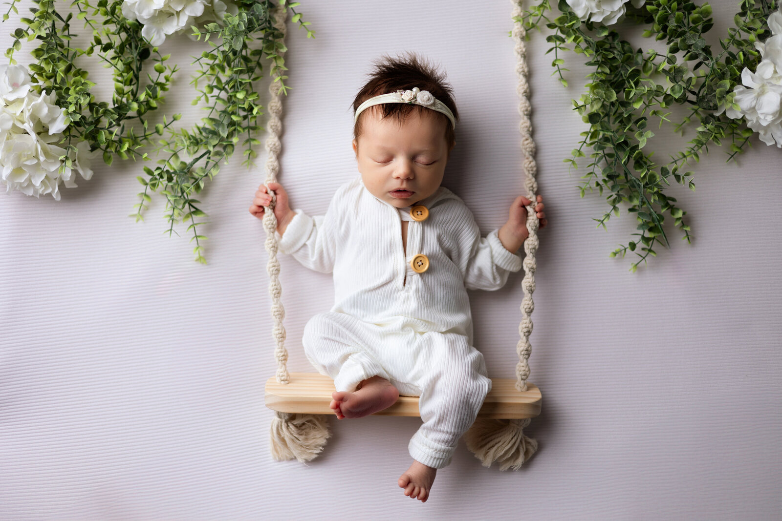 Newborn baby girl dressed in a white outfit posed on a macramé wooden swing