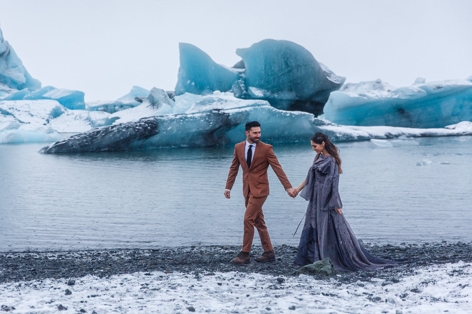 A bride and groom walk along the shores of a glacier lagoon at their winter Iceland elopement.