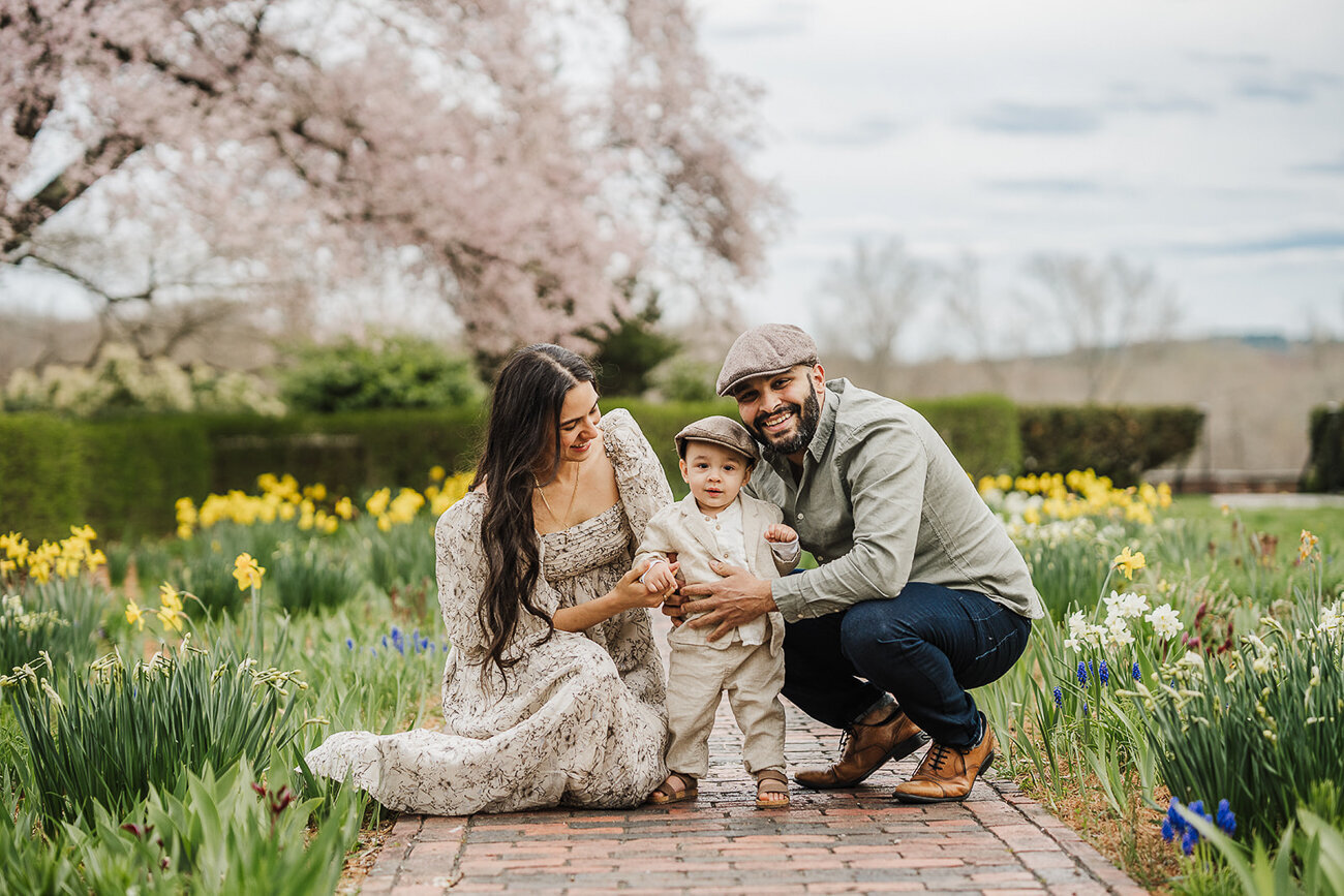 family stands in a garden full of spring flowers with toddler son