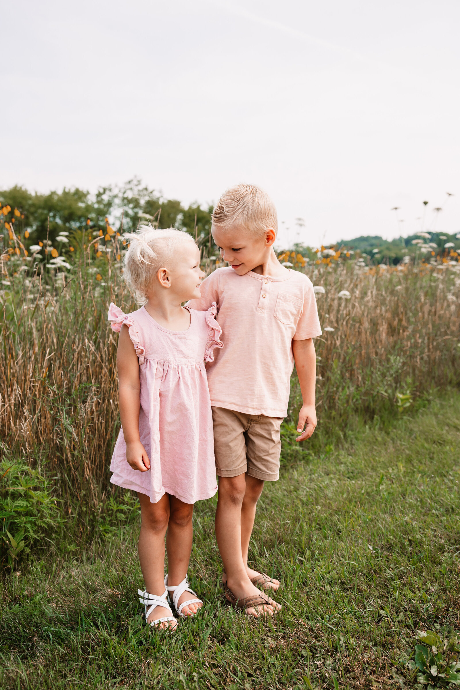 Smiling toddler boy in a pink shirt stands hugging his younger sister in a pink dress in a trail lined with wildflowers for a Milwaukee Family Photographer