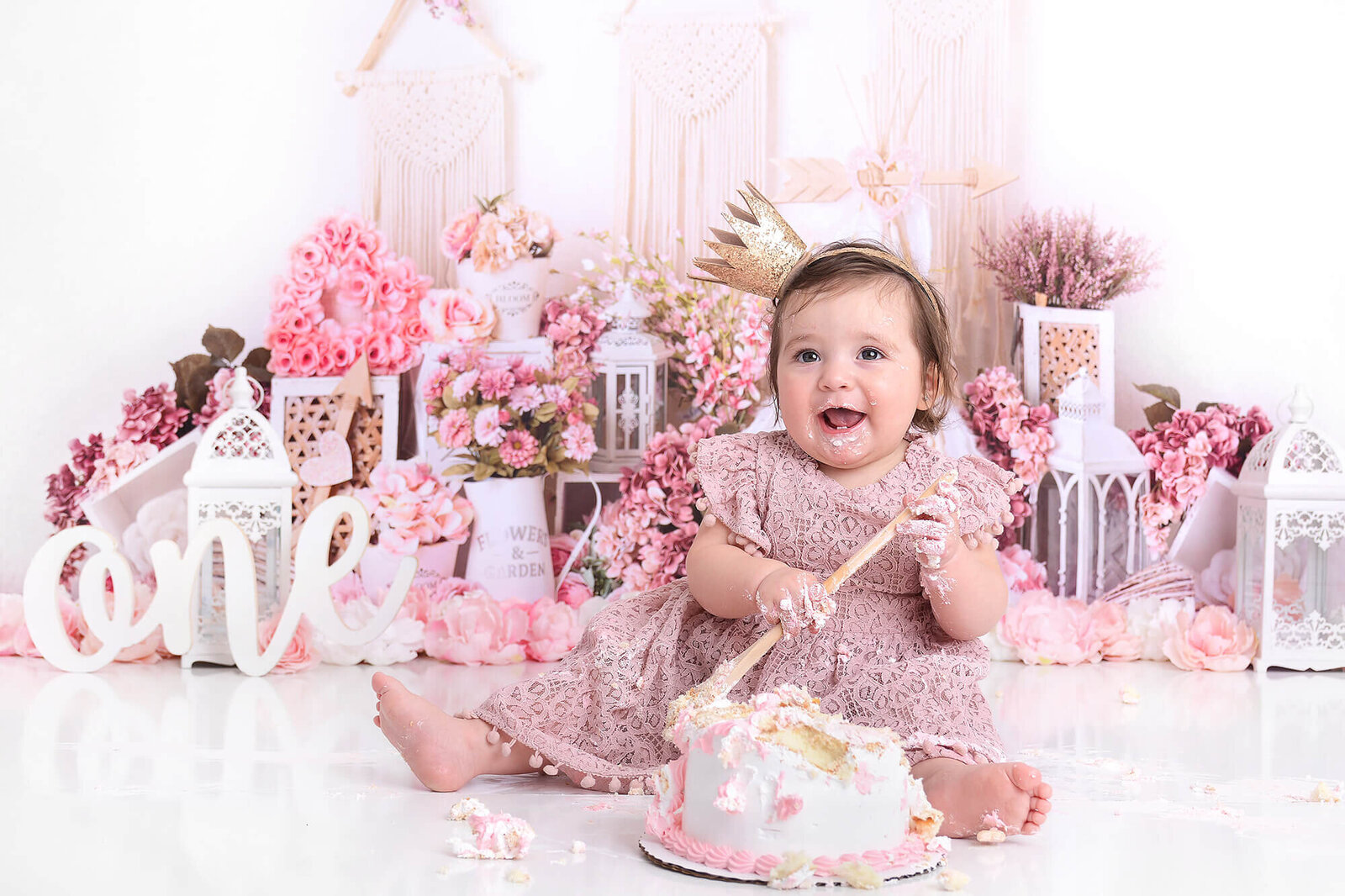girl smiles at the camera at her cake smash photography shoot with pink flowers behind her