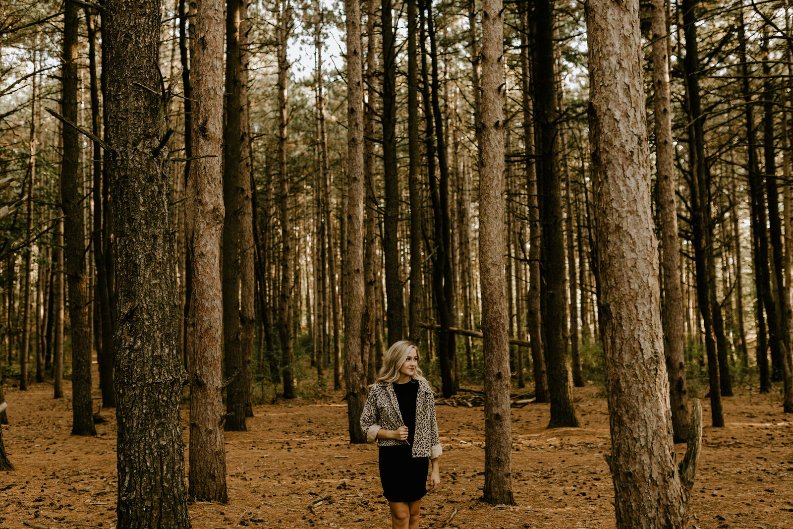 A girl holding her jacket and looking to the side while she stands in the middle of a big forest