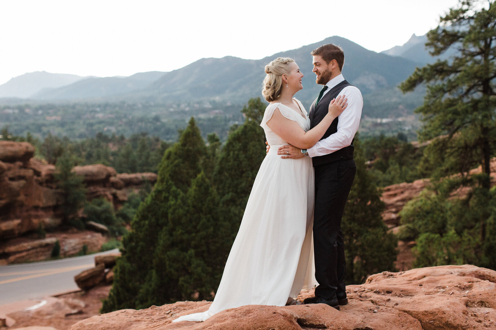 Portrait of a bride and groom posing on top of a rock formation after their wedding ceremony at the Garden of the Gods in Colorado Springs, Colorado.  The bride on the left is wearing a short sleeve, long, elegant, white dress while the man on the right is wearing a three piece suit with a tie but without the jacket. The Rocky Mountains and many trees can be seen spanning out behind them.