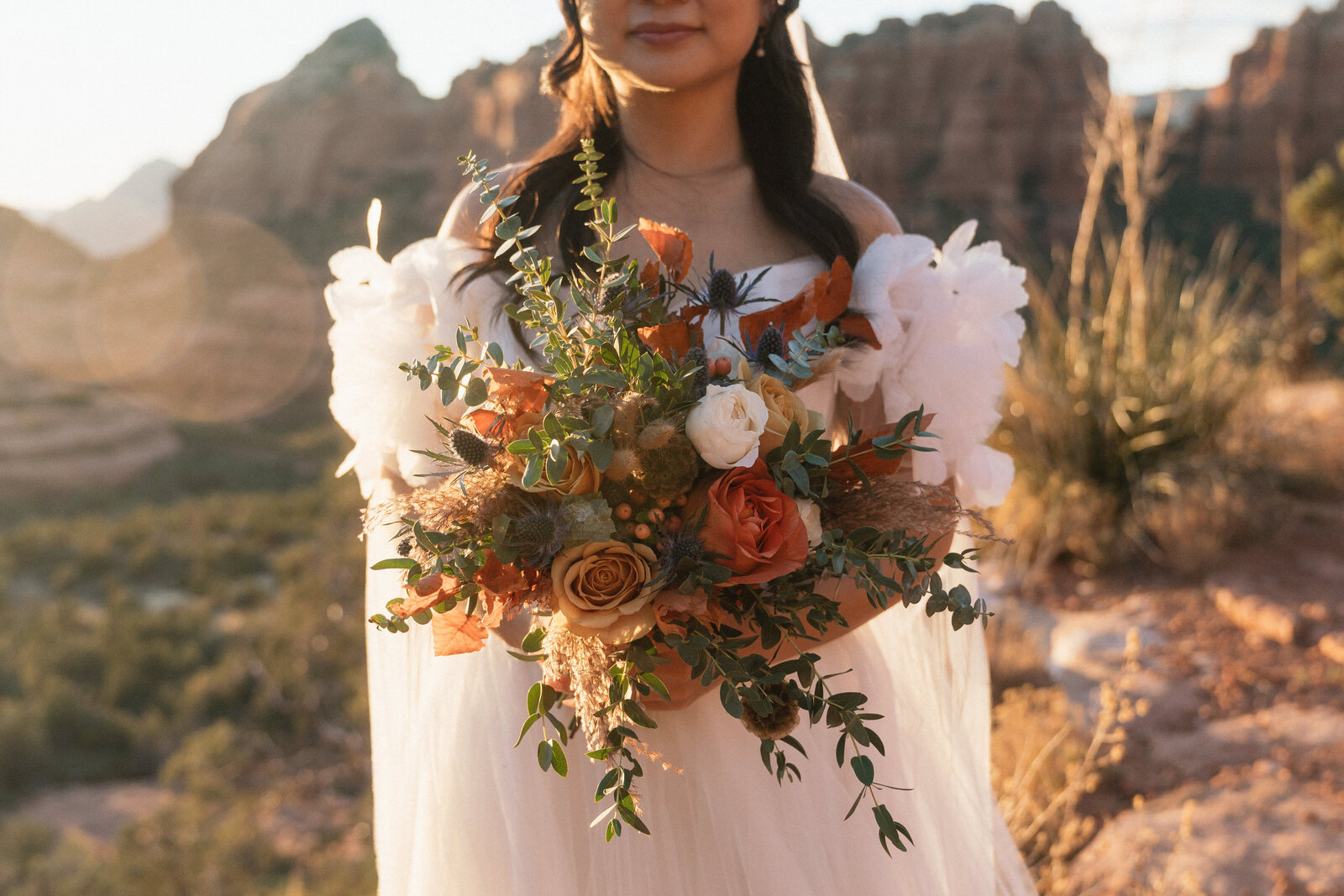 Close-up of a bride holding a warm-toned floral bouquet with roses, eucalyptus, and thistle during golden hour at Merry Go Round Rock in Sedona, Arizona, with red rock cliffs softly glowing in the background.