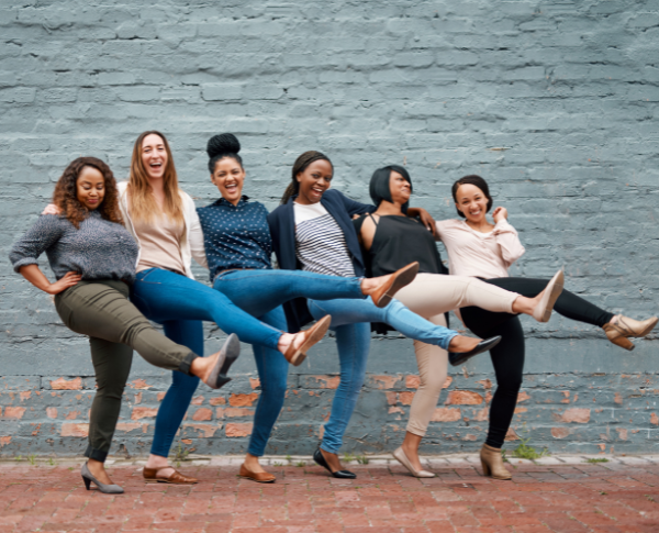 six women standing beside each other hugging and kicking legs forward while smiling