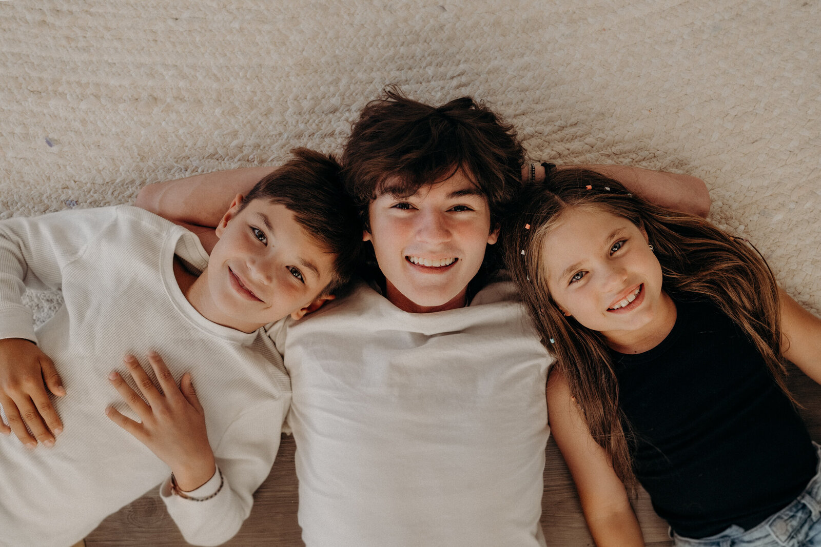 Three siblings lay on the floor, smiling up towards the audience, while their heads touch together.