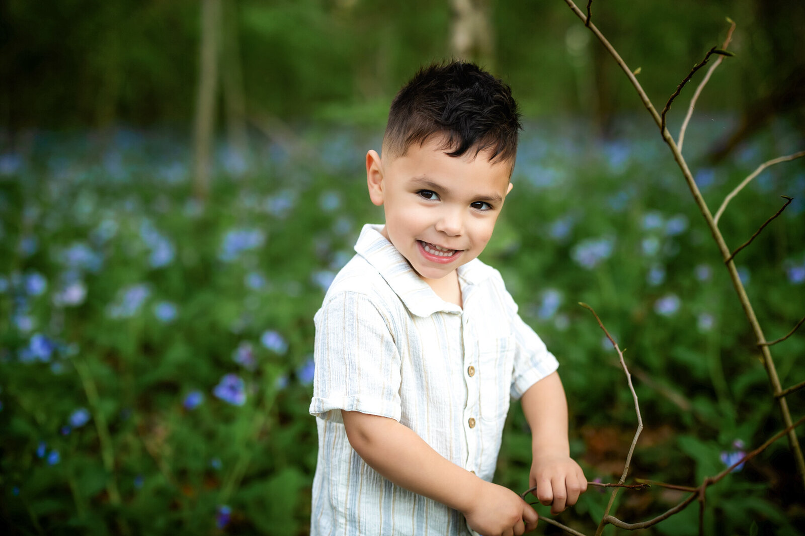 A young boy with short dark hair, wearing a light-colored shirt, smiles while standing among green foliage and purple wildflowers in a wooded area—capturing the charm of outdoor portraits by a Bloomington family photographer.