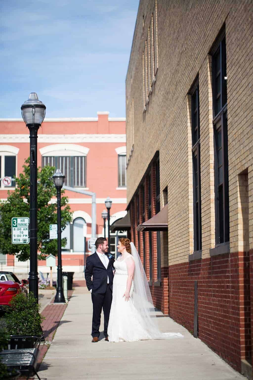 A bride in a white gown and veil stands on a sunny sidewalk next to a groom in a dark suit. They are smiling at each other, surrounded by brick buildings and street lamps.