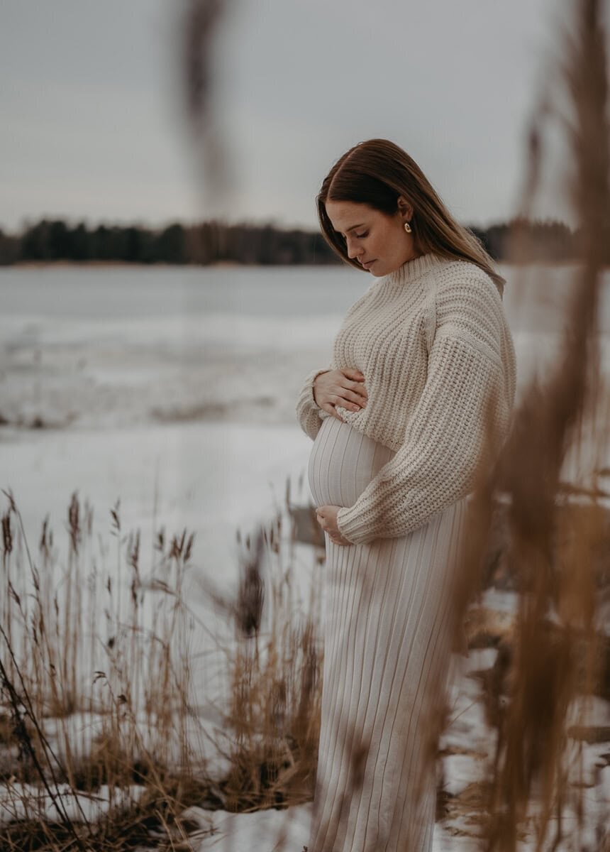 Blivande mamma står i vinterlandskap i stickade kläder och håller om sin gravida mage.