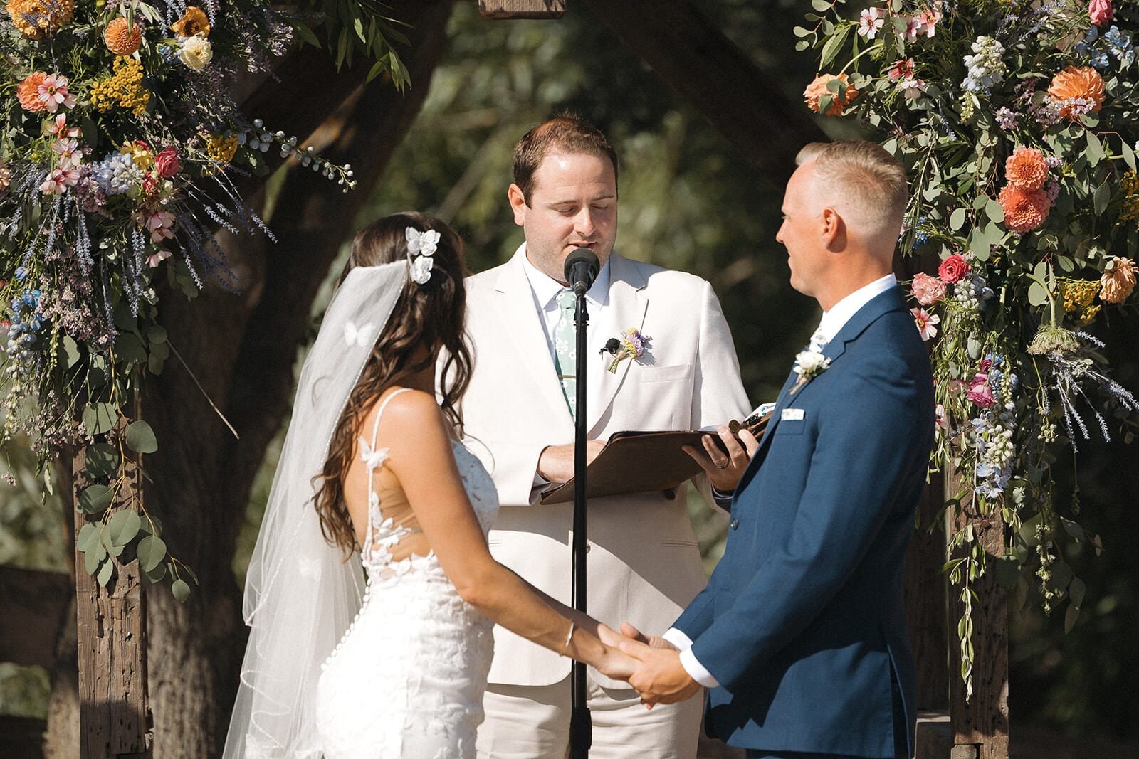 Bride and groom holding hands during their vows under a floral arbor