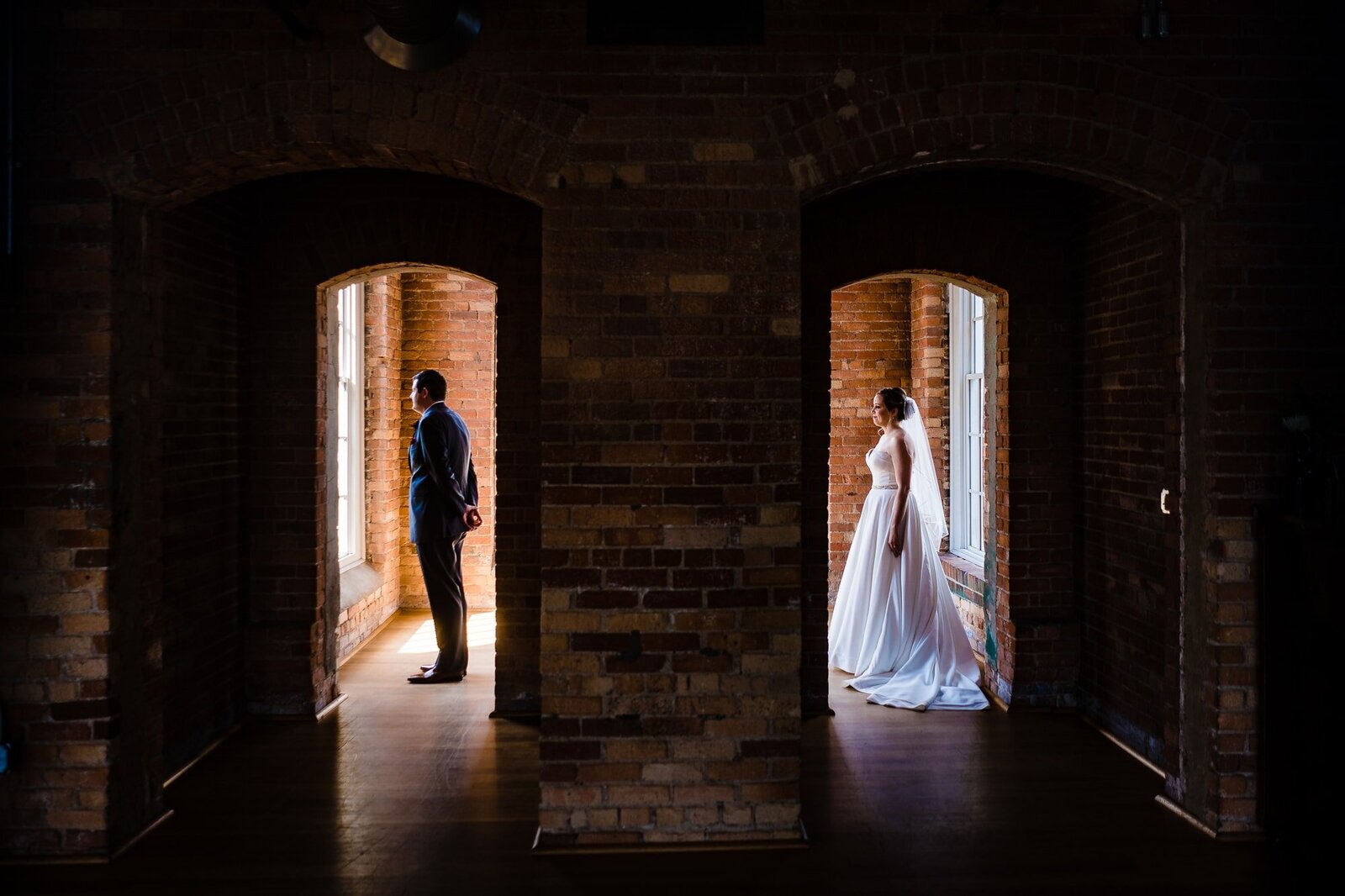 Bride stands behind groom the moment before their first look. He is seen through an archway on the left, and she is seen through another archway on the right
